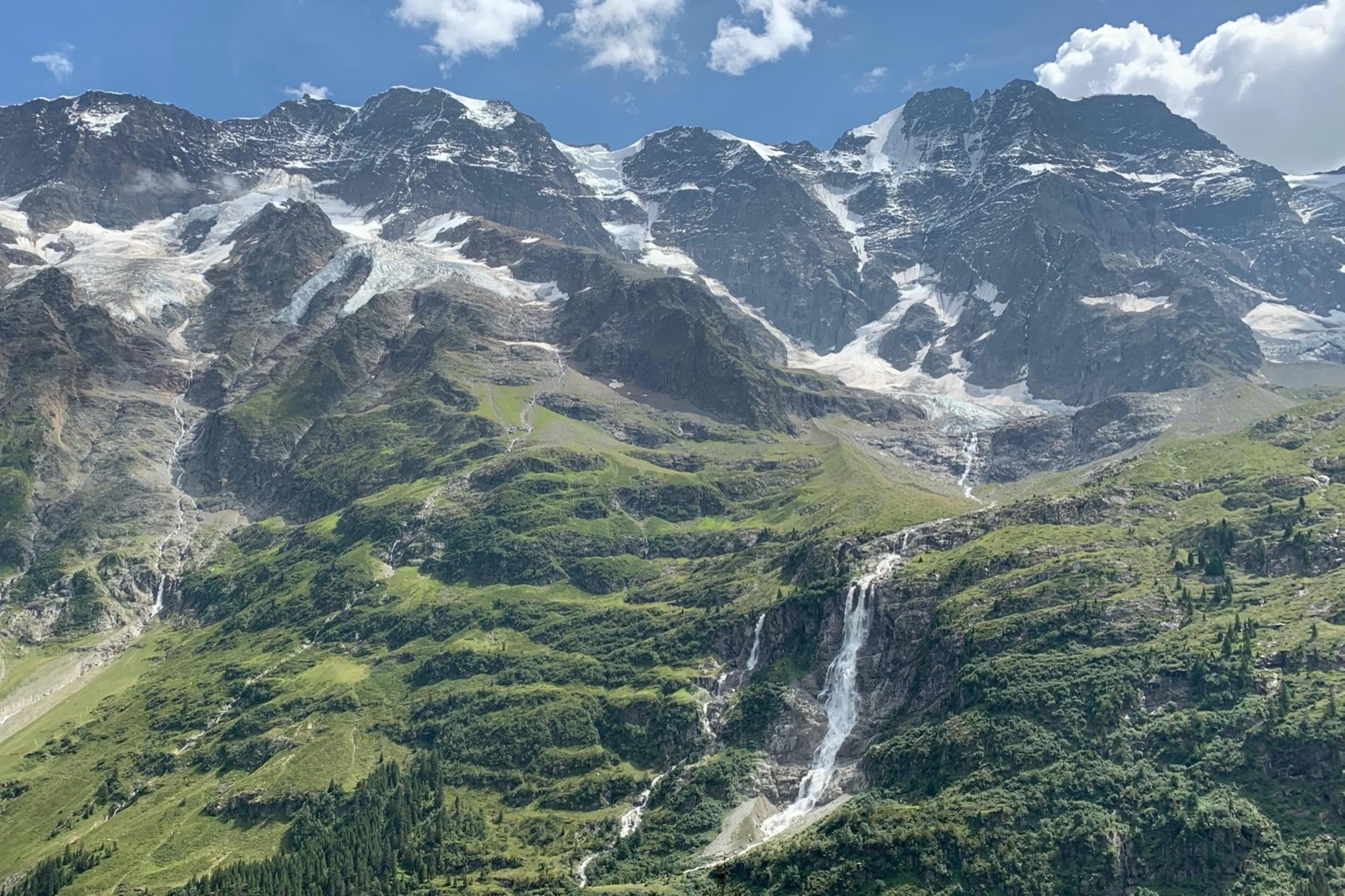 Schneebedeckte Berge ragen in den blauen Himmel. Die Vegetation verändert sich talabwärts von Gestein zu Wald- und Grünflächen. Mittig befindet sich ein Wasserfall.