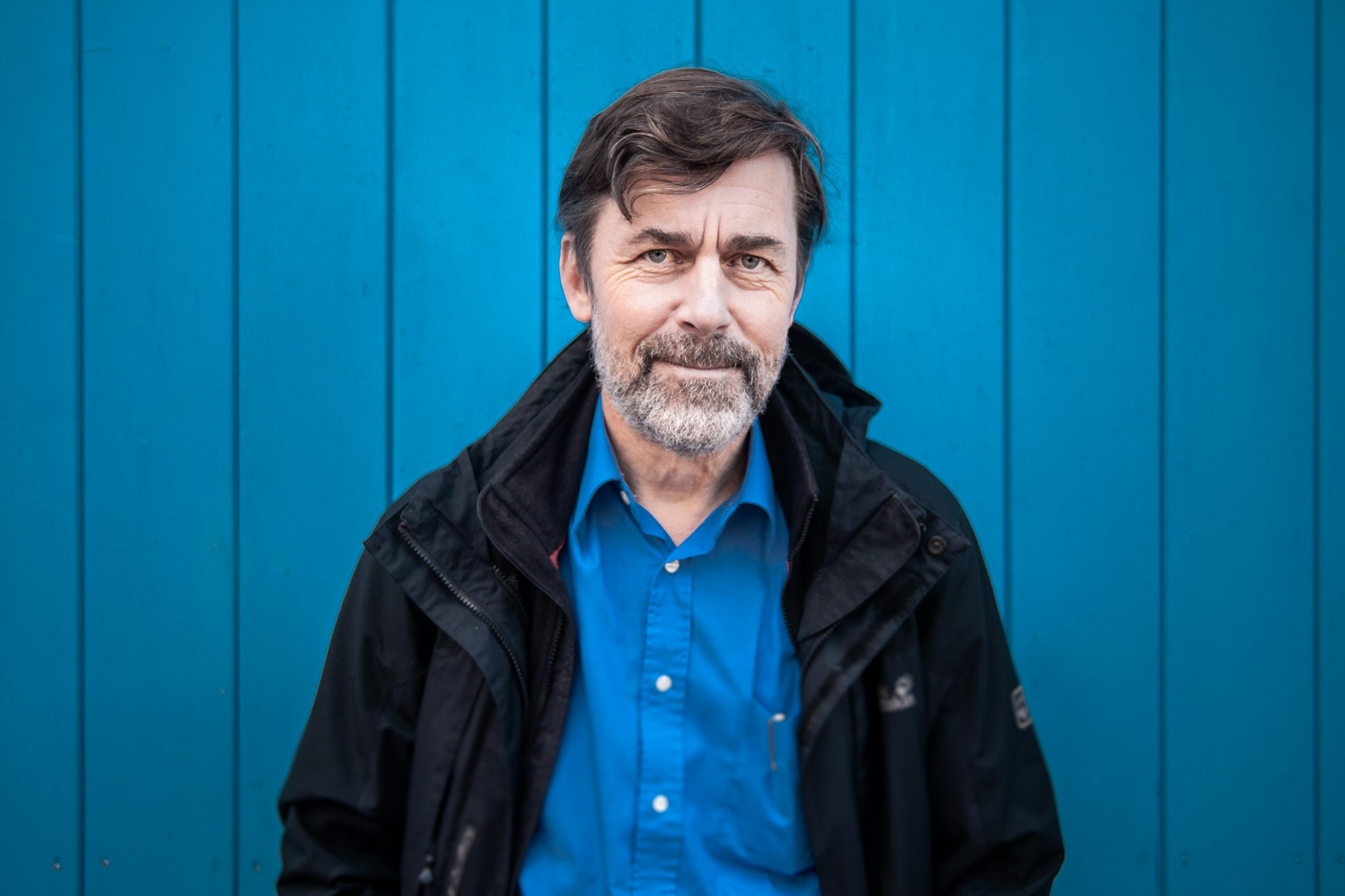 Fotoportrait eines Mannes, der ein blaues Hemd und eine dunkle Wetterjacke trägt, vor einer blaugestrichenen Wand.