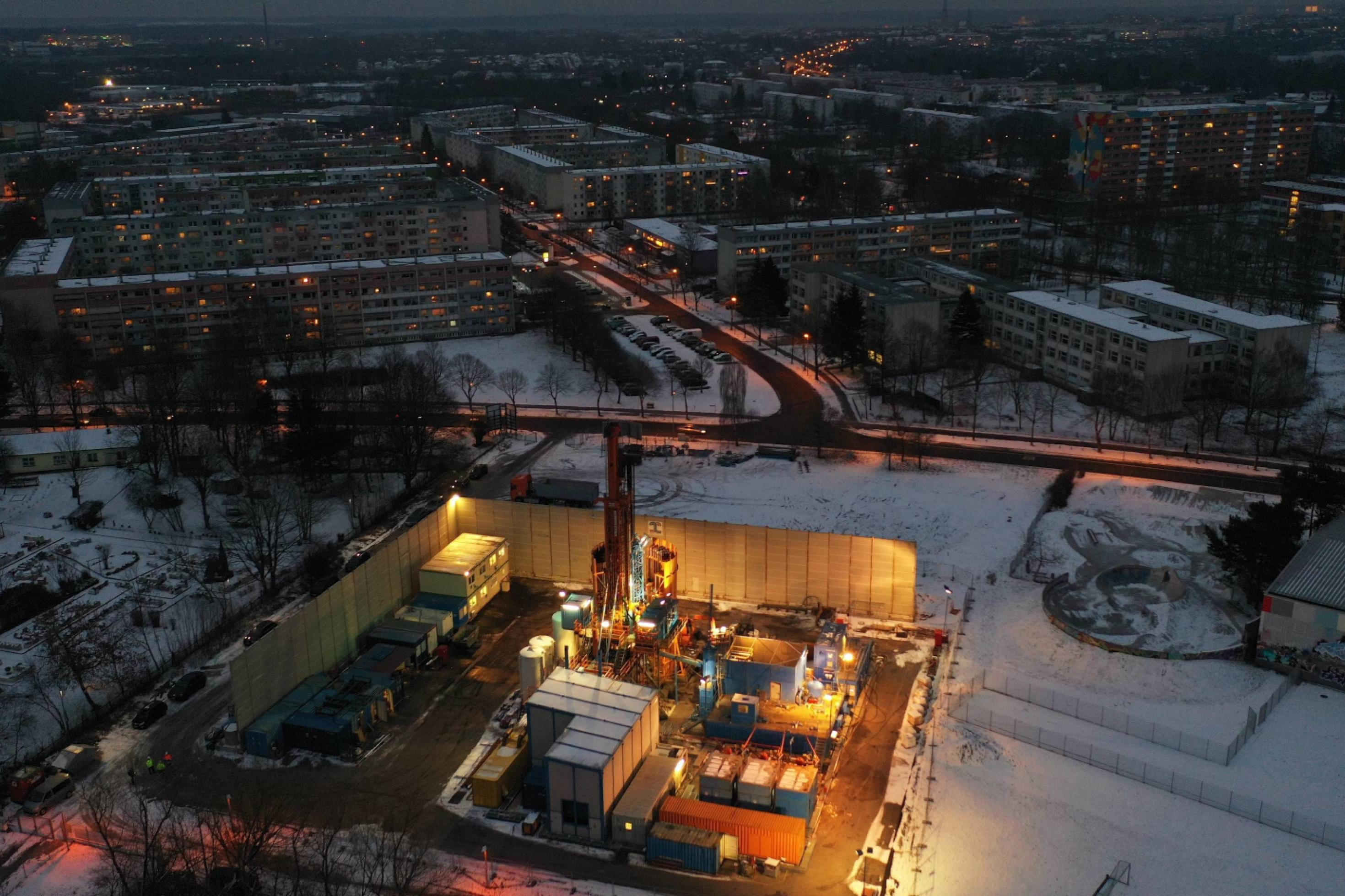 Luftbild einer verschneiten Landschaft im Dämmerlicht, in der Ferne Plattenbauten und Straßen, im Vordergrund ein gut angestrahlter Bohrplatz mit Bohrturm, Baucontainern.