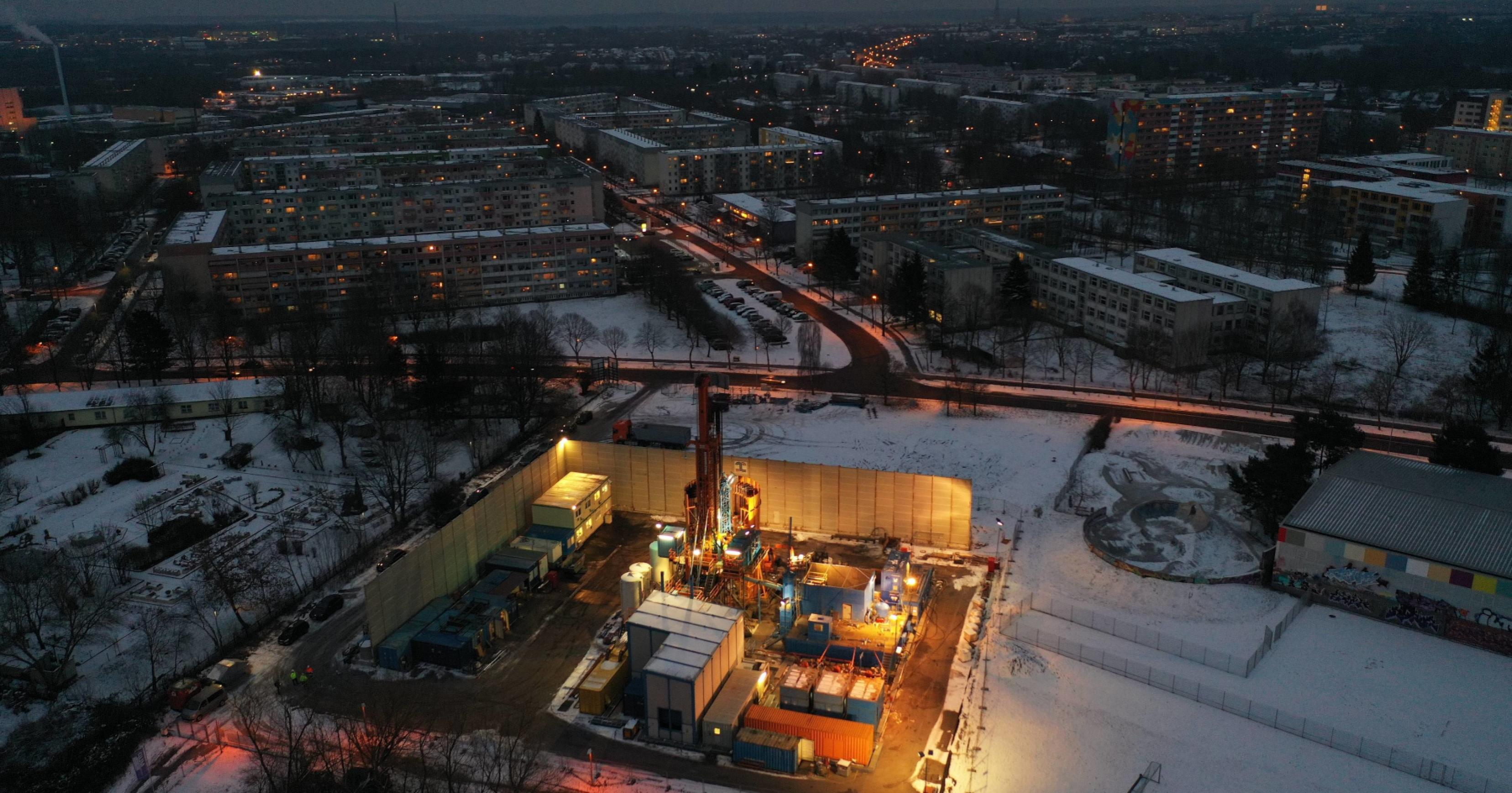 Luftbild einer verschneiten Landschaft im Dämmerlicht, in der Ferne Plattenbauten und Straßen, im Vordergrund ein gut angestrahlter Bohrplatz mit Bohrturm, Baucontainern.