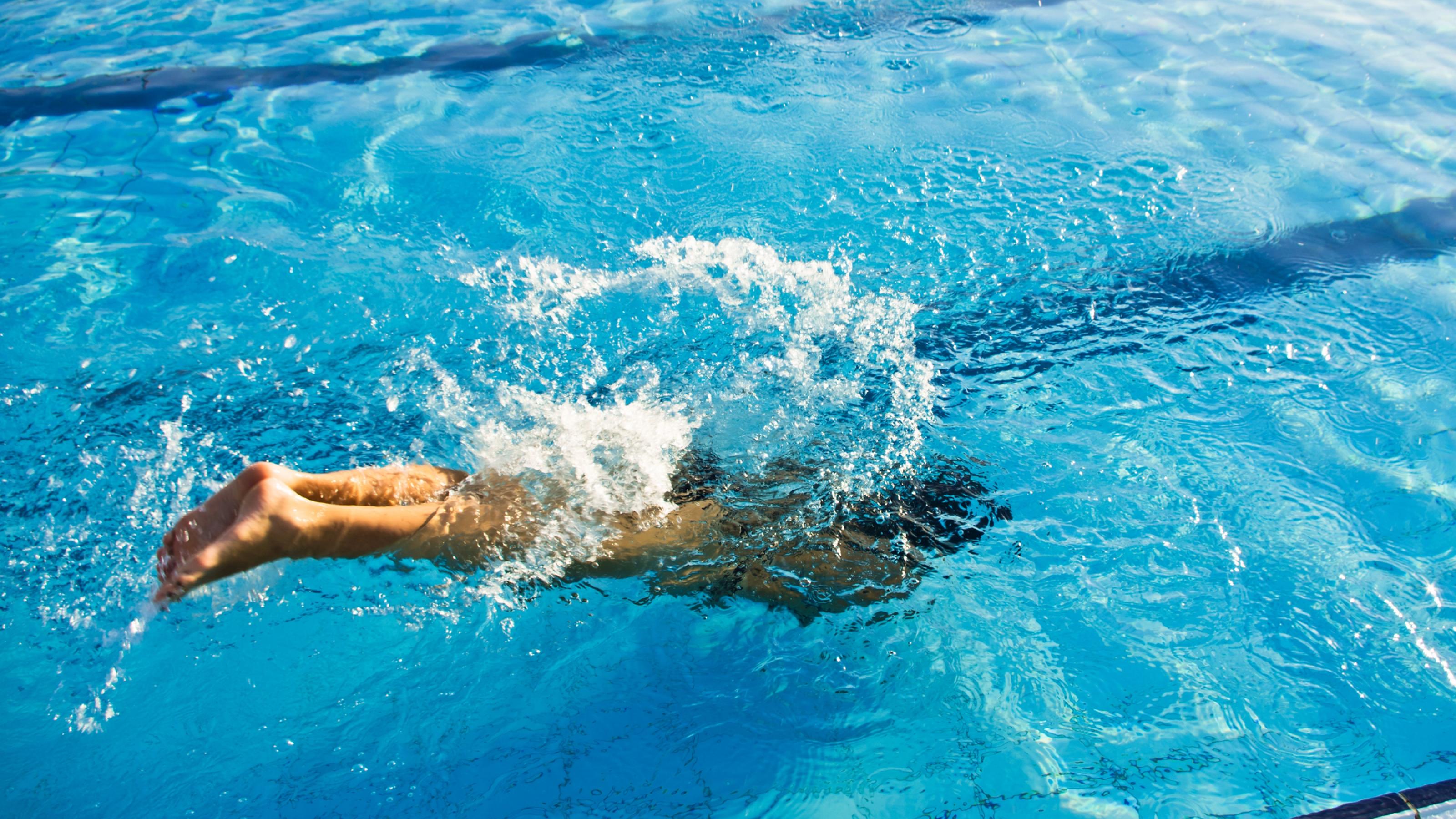 Ein Mensch taucht kopfüber in ein hellblaues Freibadbecken. Wasser spritzt auf, die Beine ragen noch aus dem Wasser. Die Sonne scheint auf der Wasseroberfläche.