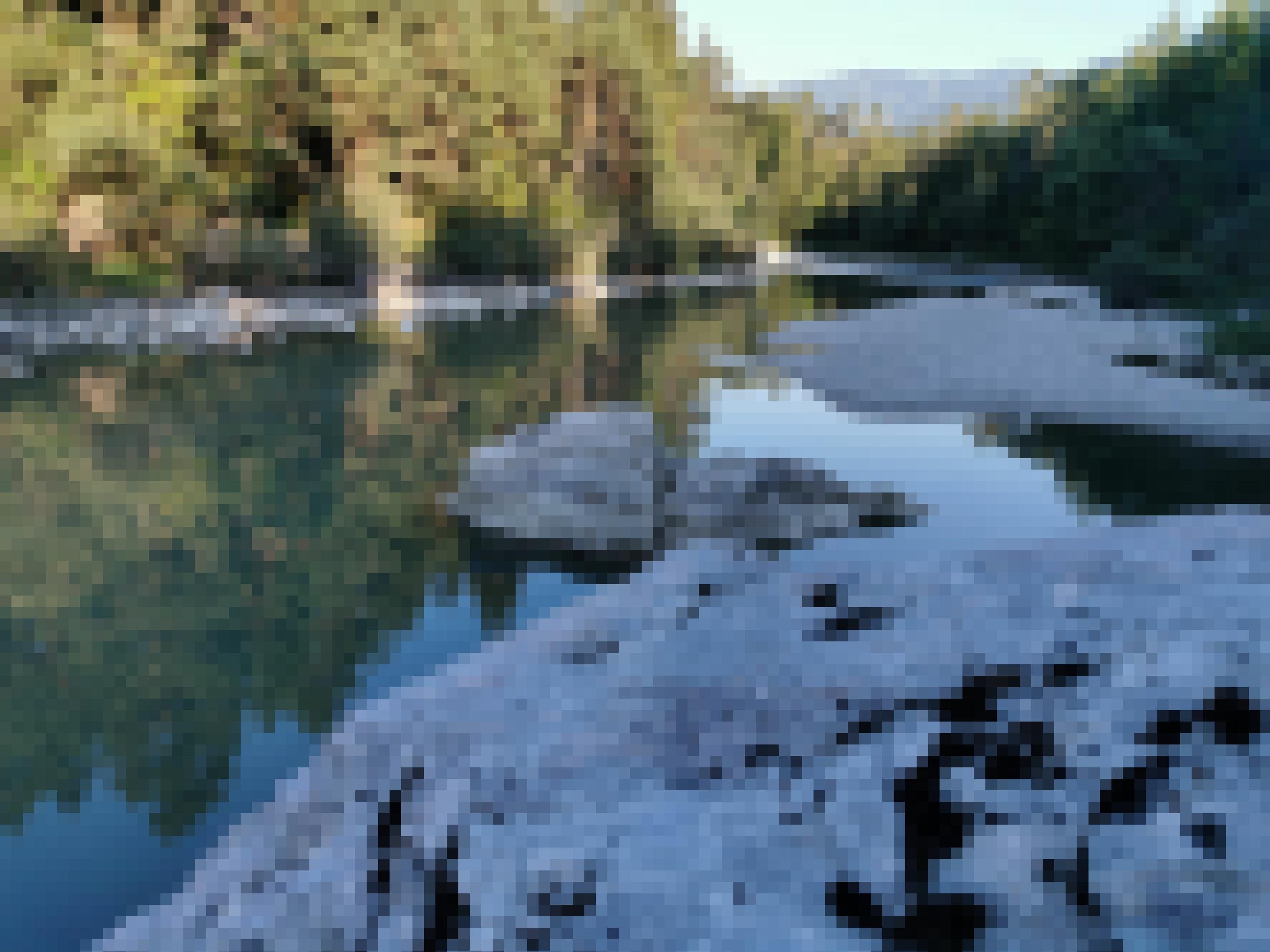 Nadelwald, dunkelgrünes Flusswasser, Schotterufer und große Steine.