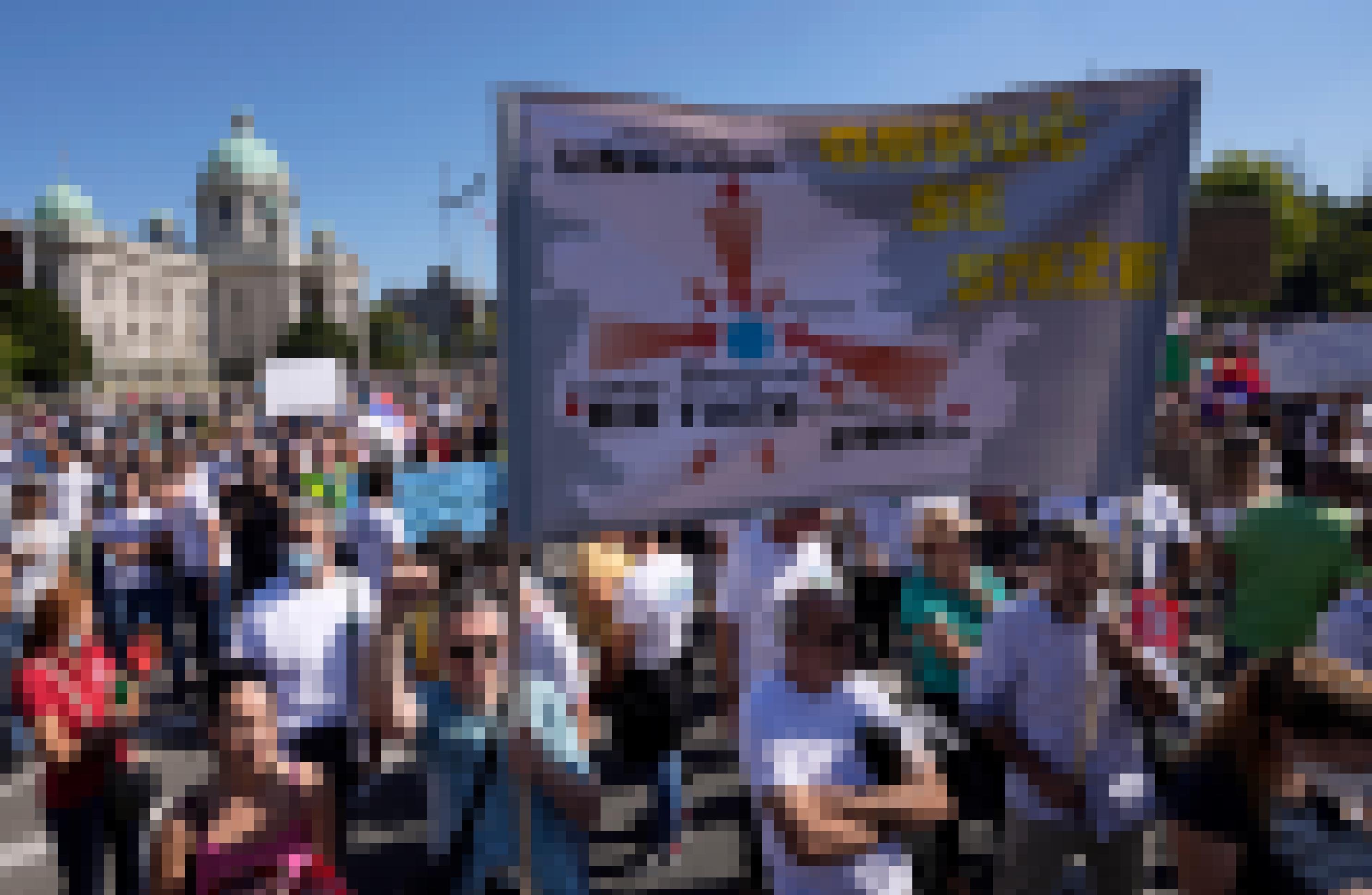 Menschen demonstrieren auf einer sonnigen Straße. Ein Mann erhebt die Faust. Ein professionell gestaltetes Plakat zeigt Pfeile, die auf Belgrad weisen.