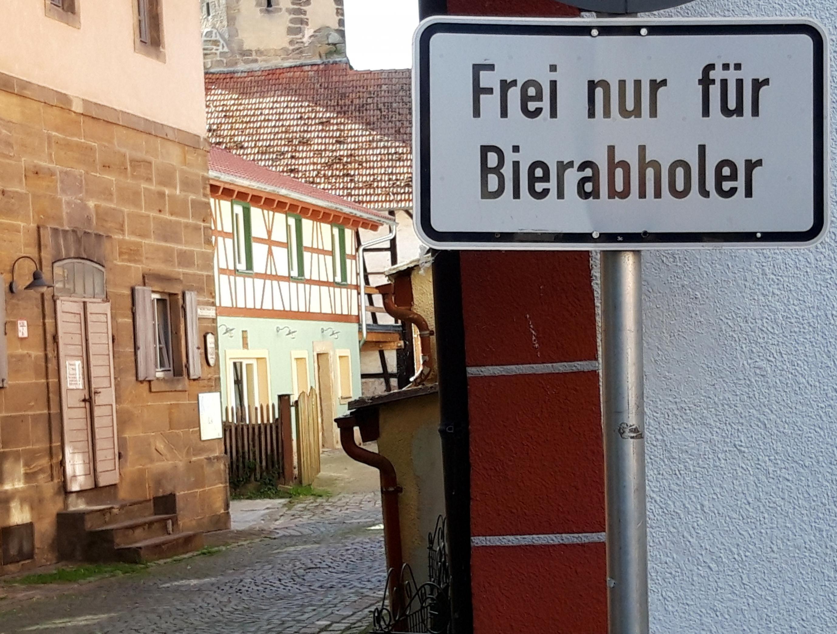 Vor einer im Abendlicht schimmernden Gasse mit Fachwerkgebäuden über Kopfsteinpflaster steht das Straßenschild mit der Aufschrift „Frei nur für Bierabholer“.