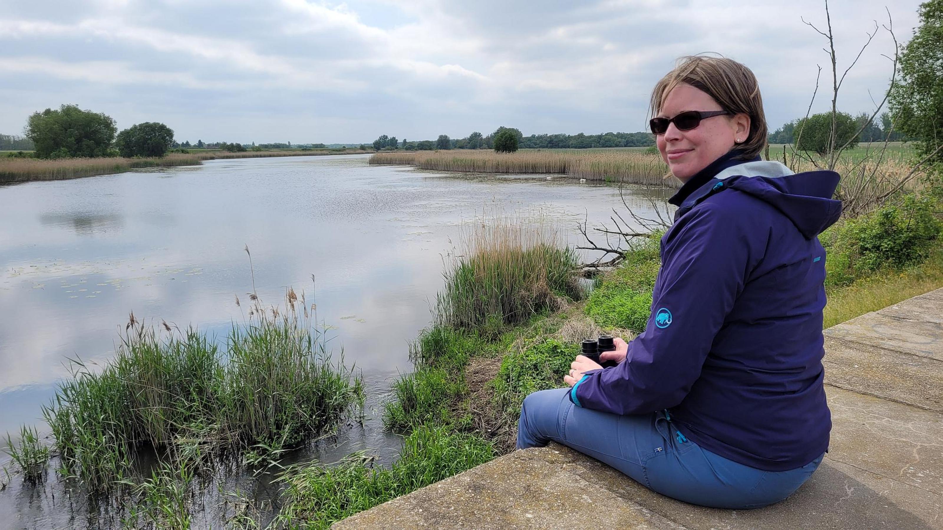 Frau in blauem Anorak auf einem Holzsteg, im Hintergrund schilfgesäumte Wasserfläche