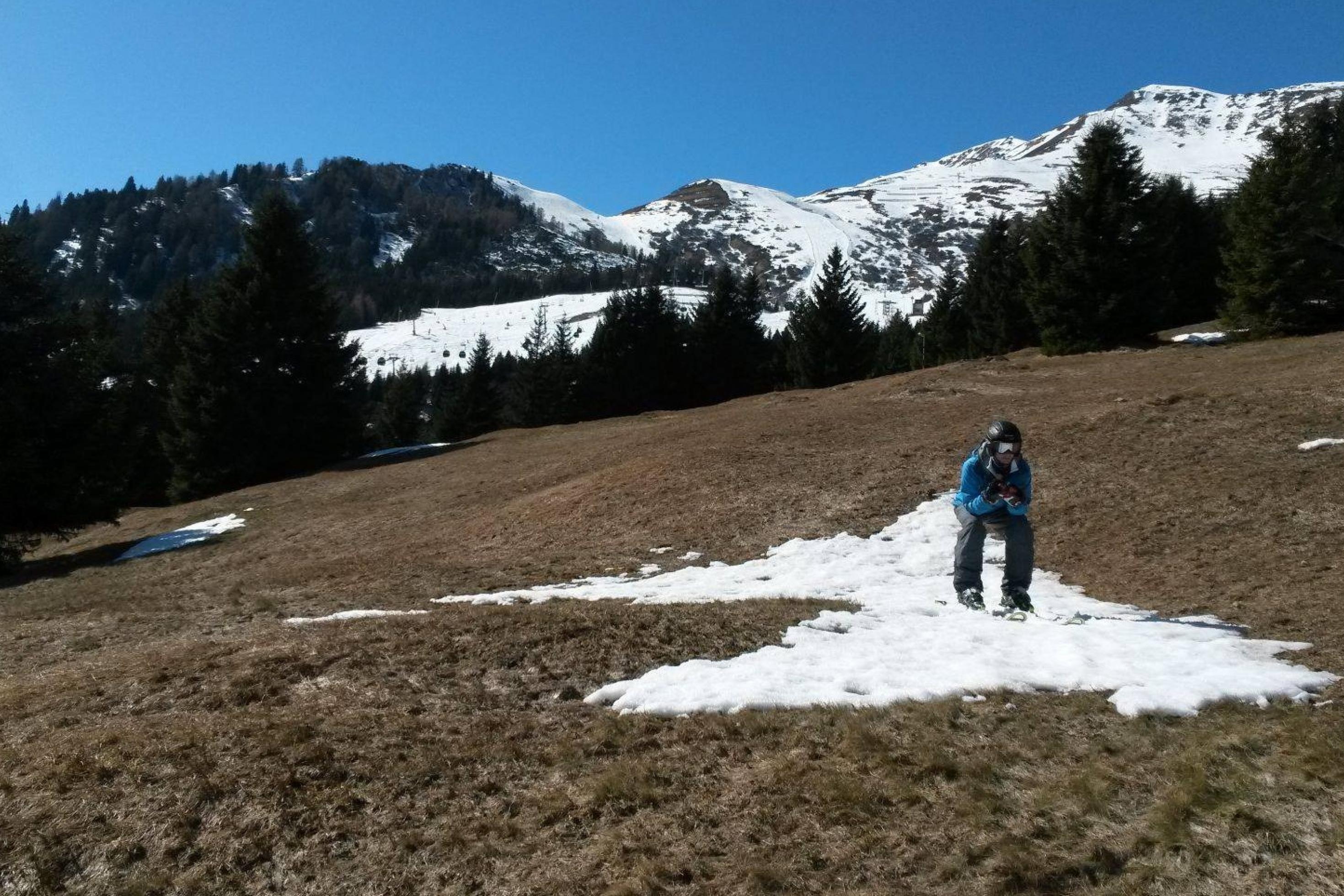 In einer braunen Bergwelt steht ein einsamer Skifahrer in voller Montur auf einem winzigen Schneefeld. Die Abfahrt scheint nur wenige Meter lang zu sein.