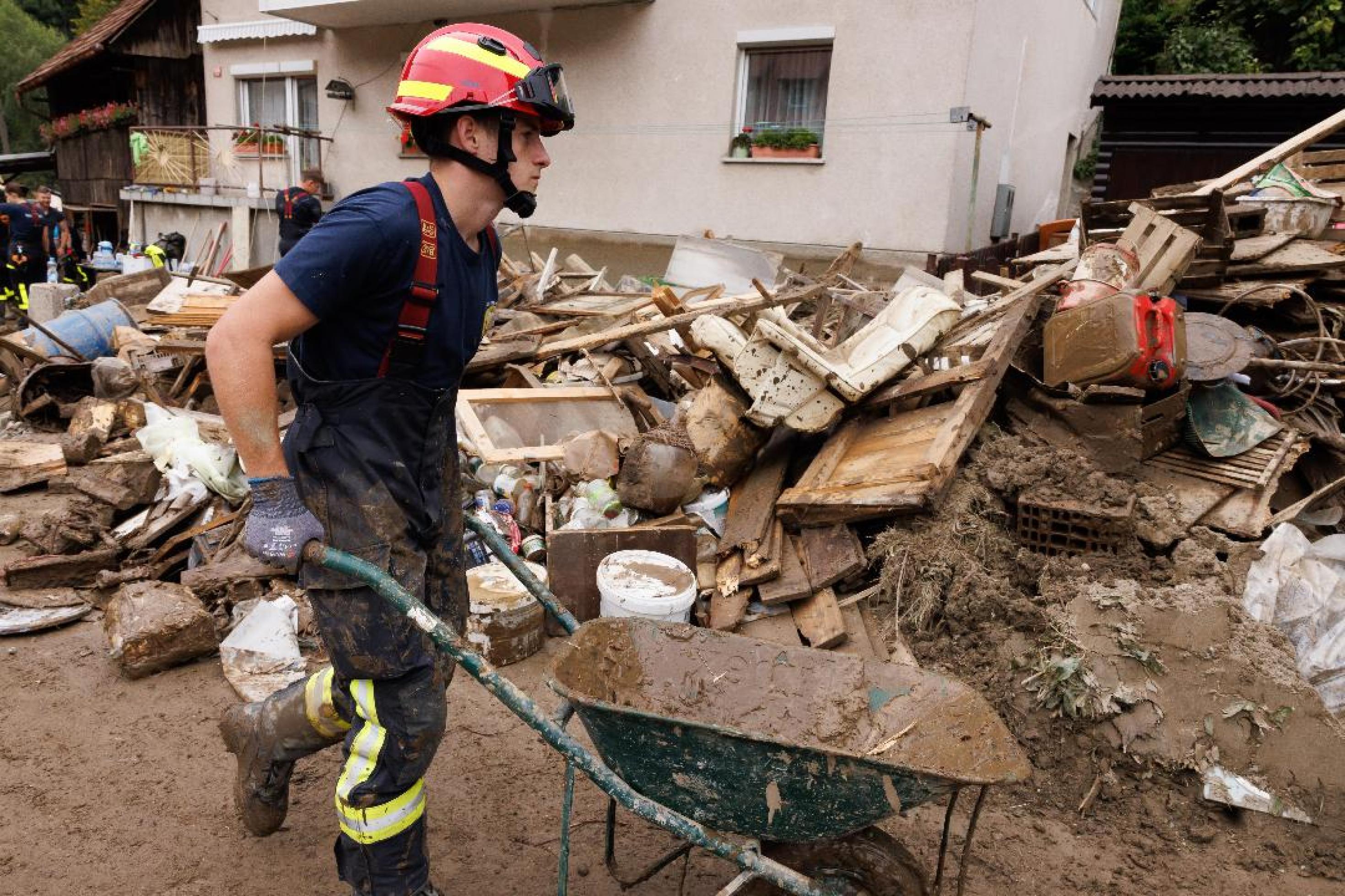 Ein junger Feuerwehrmann schiebt eine Schubkarre neben einem großen Haufen von zerstörtem Hausrat. Die Straße und der Müllberg sind von Schlamm überzogen.