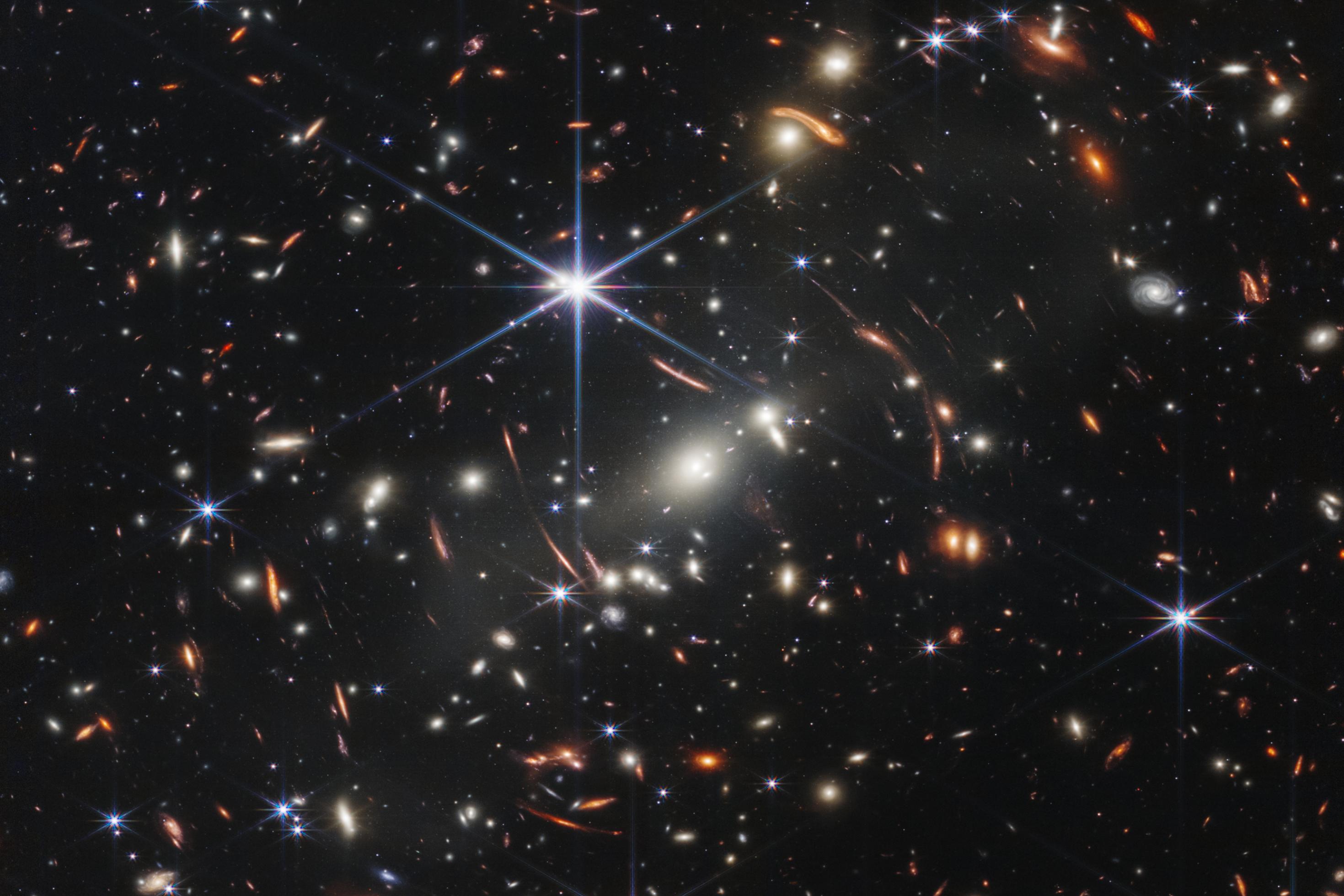 In dieser ersten Aufnahme des James Webb Space Telescope sind zahlreiche Galaxien zu sehen. Die zentralen in der Bildmitte gehören eine Galaxiencluster an, der mit seiner Masse das Licht dahinter liegender Galaxien umlenkt und verzerrt. So erschienen um den zentralen Cluster herum gebogene Objekte, Galaxien aus dem frühesten Universums.