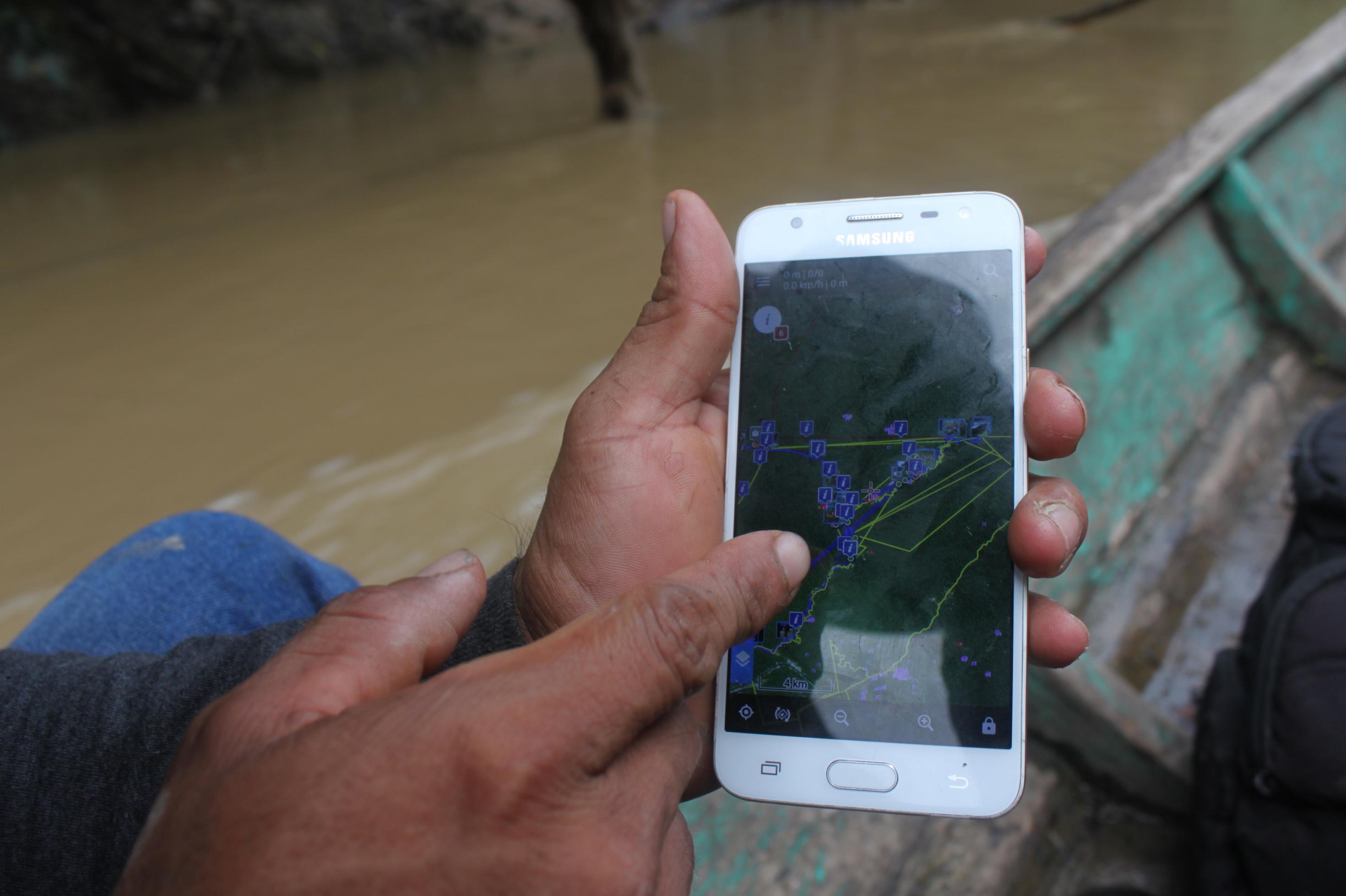 Eine Hand hält ein Smartphone, die Person sitzt in einem Holzboot, das auf braunem Wasser fährt. .
