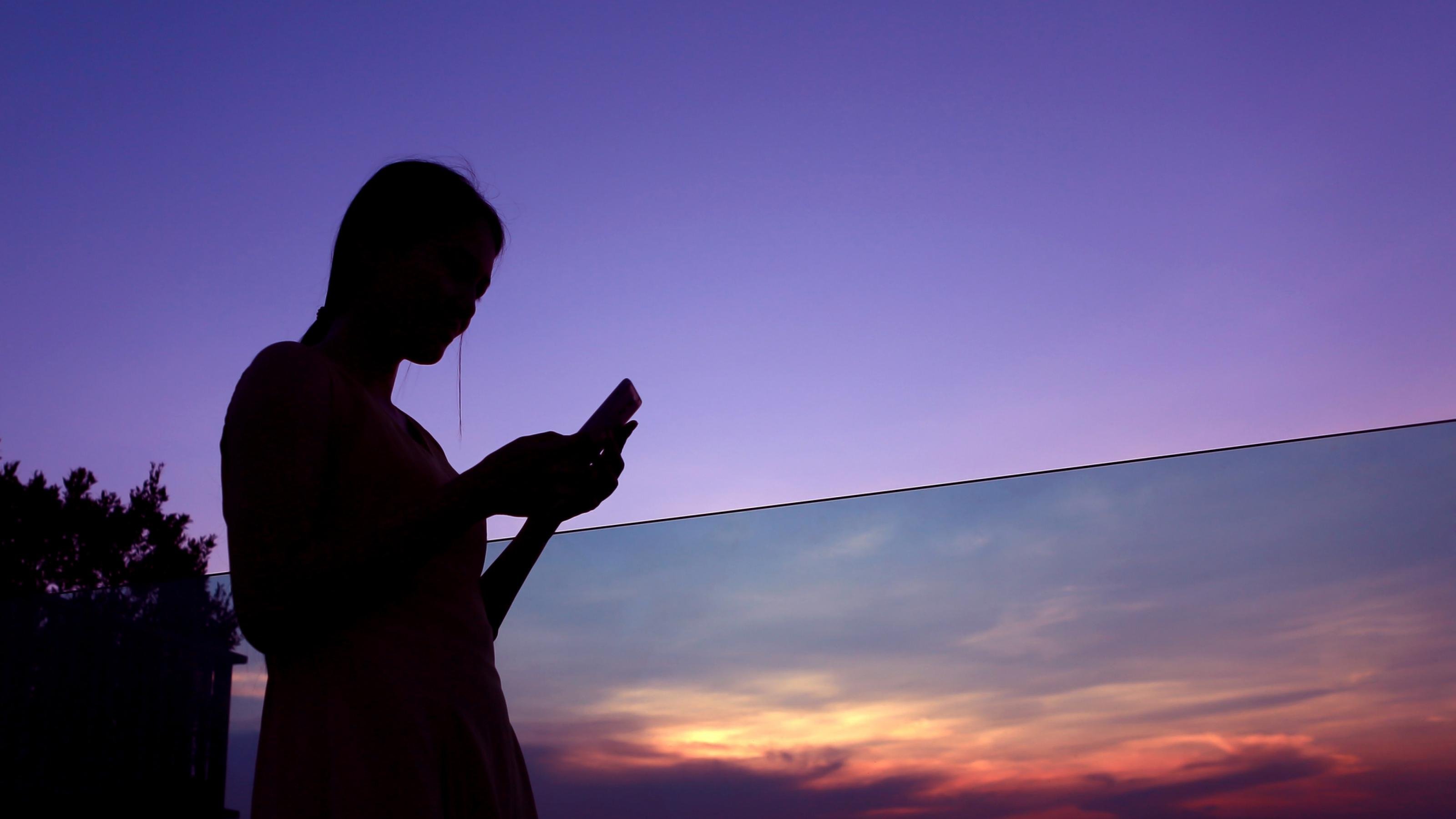 Die Silhouette einer jungen Frau vor einem lila Abendhimmel. Sie hält ein Smartphone in der Hand