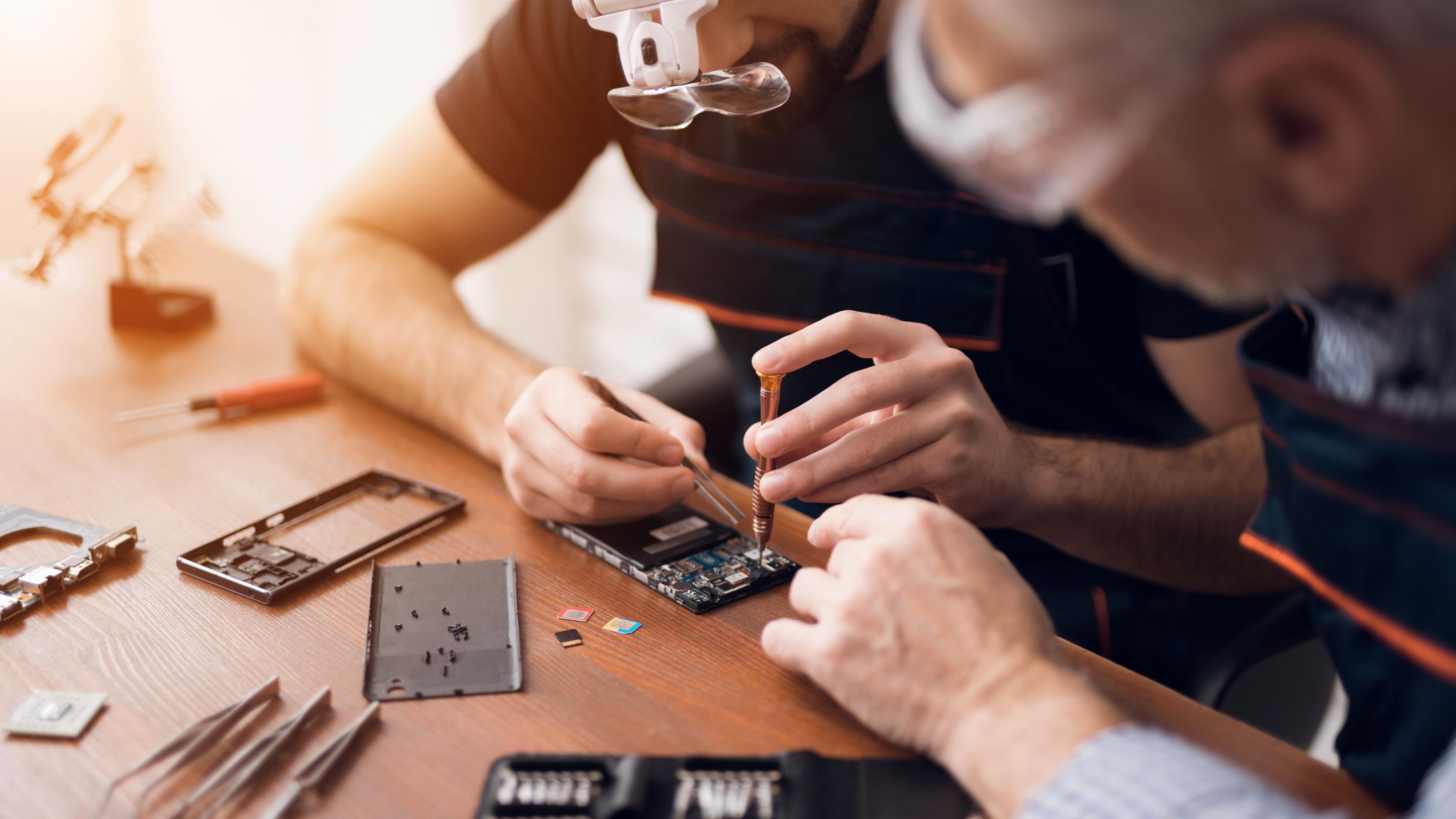 Ältere und junge Männer reparieren gemeinsam ein Mobiltelefon. Sie arbeiten in einer Werkstatt. Sie benutzen verschiedene Werkzeuge. Sie sind Reparaturspezialisten.