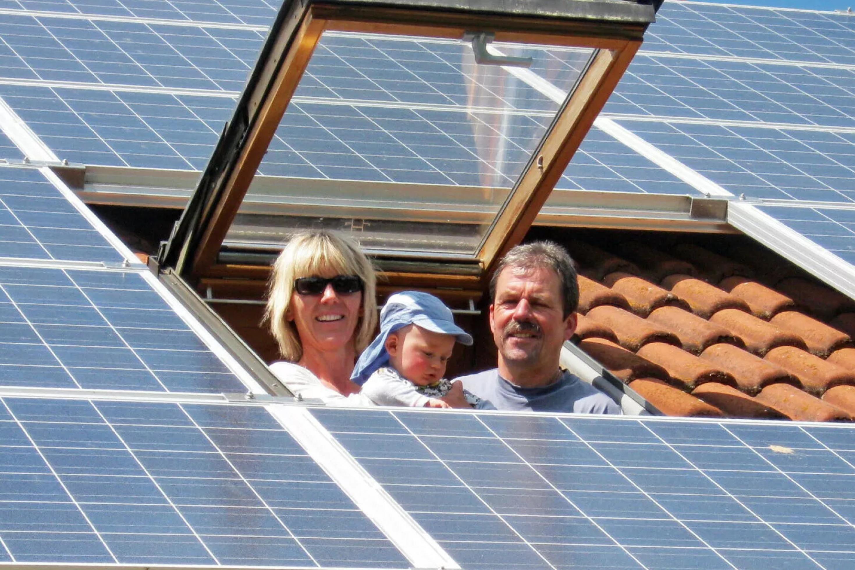 Mann, Frau und Kleinkind schauen aus einem Dachfenster, das Dach ist mit Photovoltaik-Modulen bleegt.