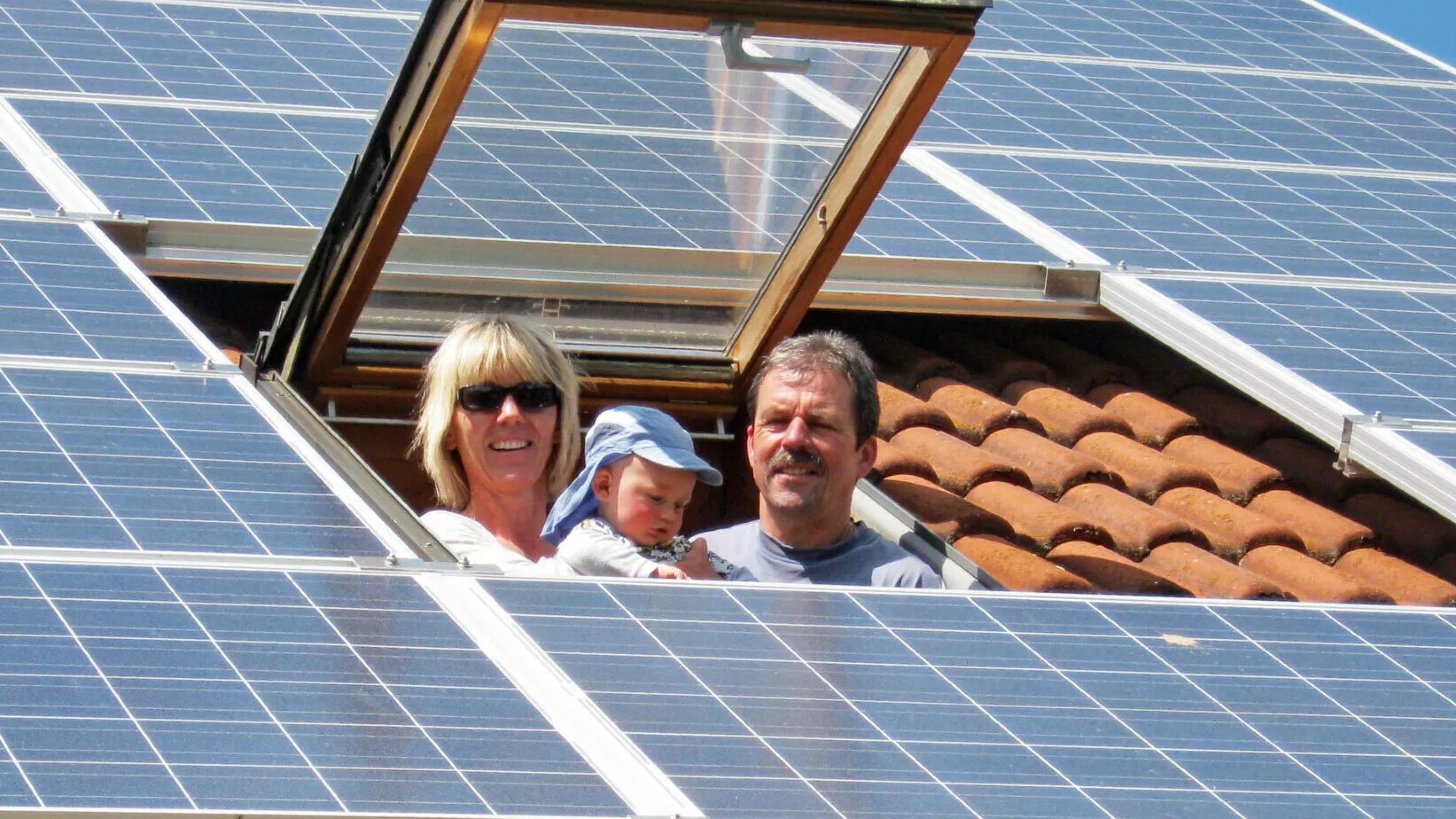 Mann, Frau und Kleinkind schauen aus einem Dachfenster, das Dach ist mit Photovoltaik-Modulen bleegt.