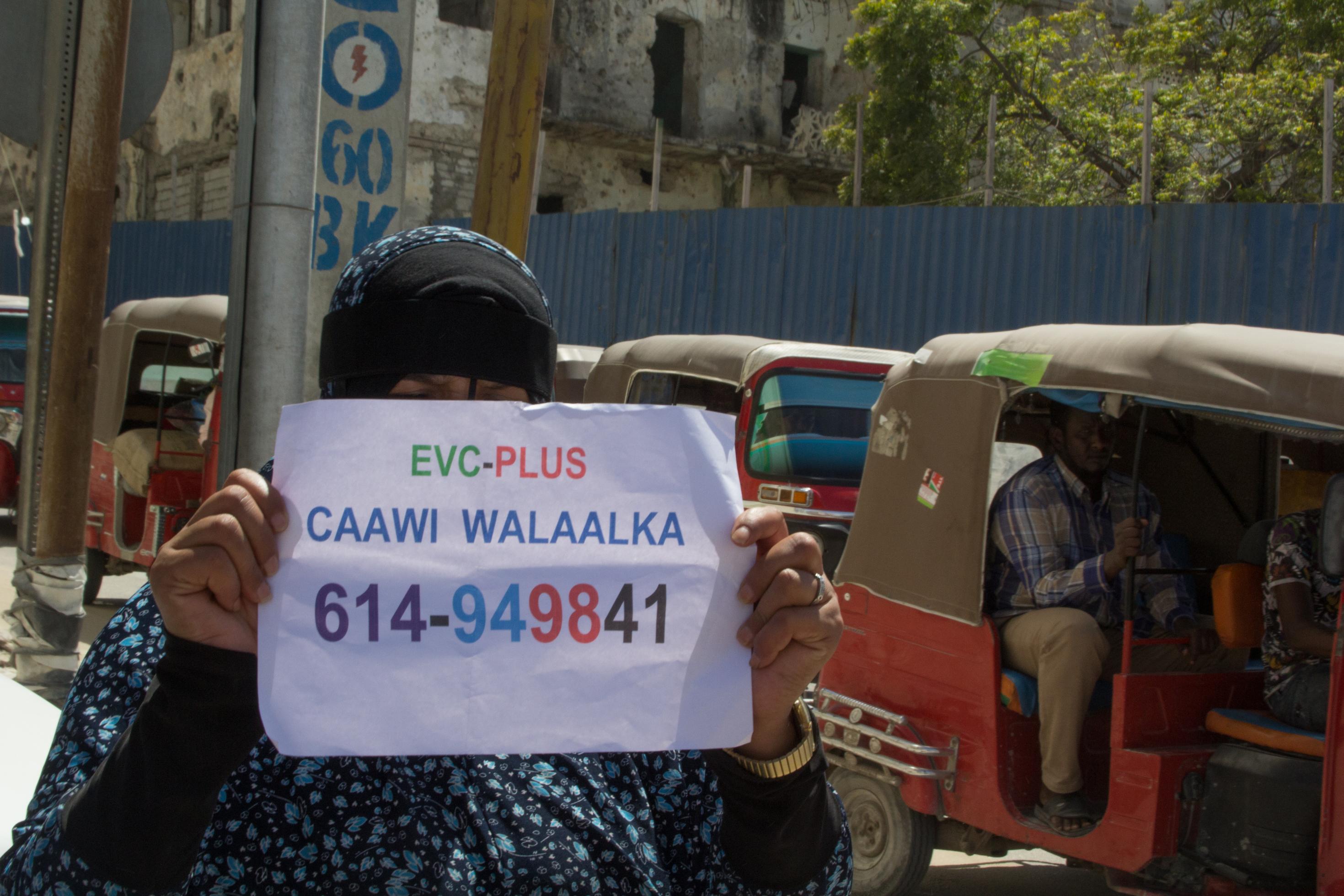 Ein Frau bettelt um e-money in Somalia 2021. Sie hält einen Zettel in der Hand mit ihrer Handynummer. Auf dem Papier steht: EVC-PLUS, CAAWI WALAALKA und eine Nummer.
