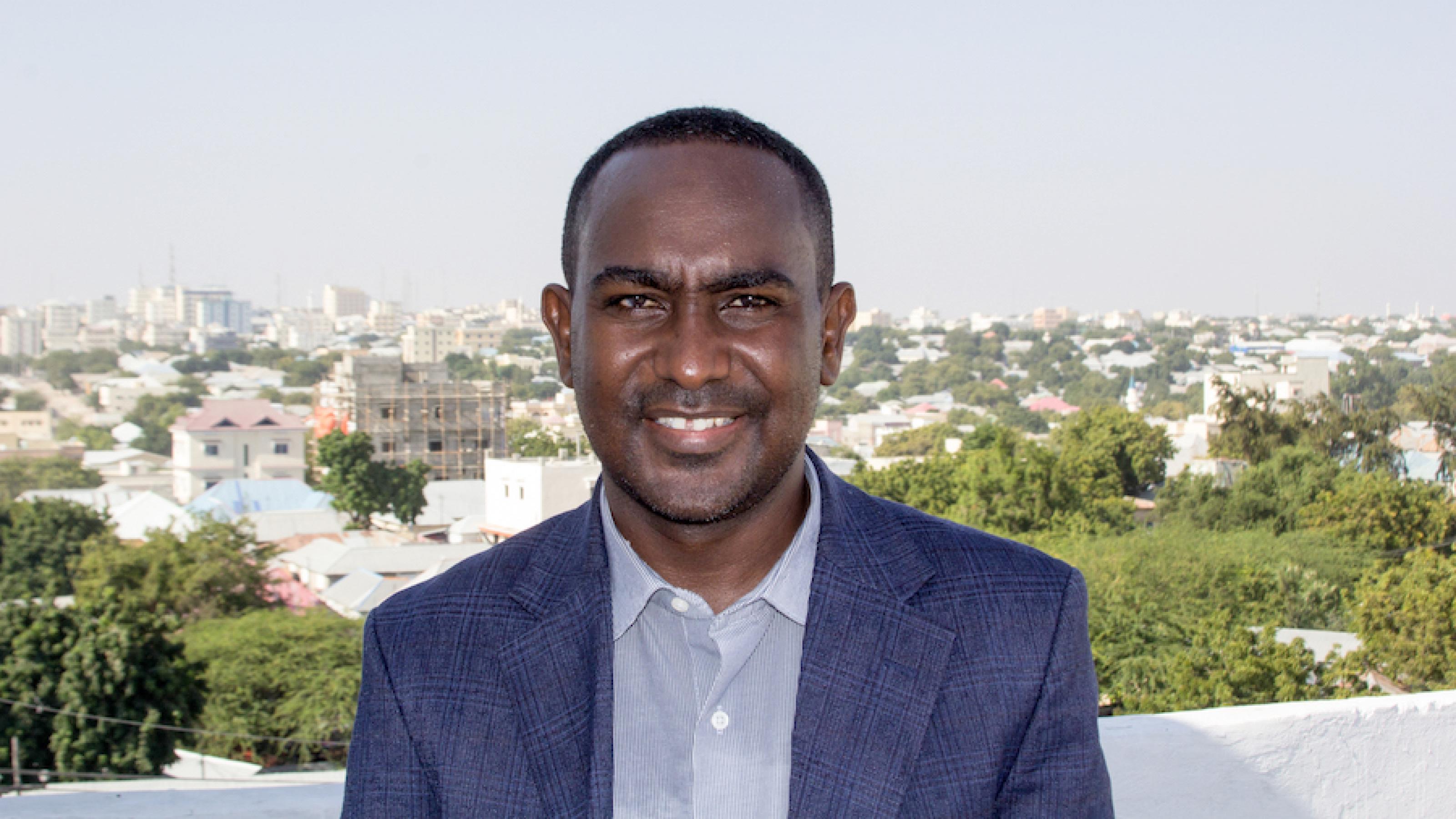 Ein Porträtfoto des Journalisten, er trägt einen Anzug, hinter ihm ist Mogadishu zu sehen