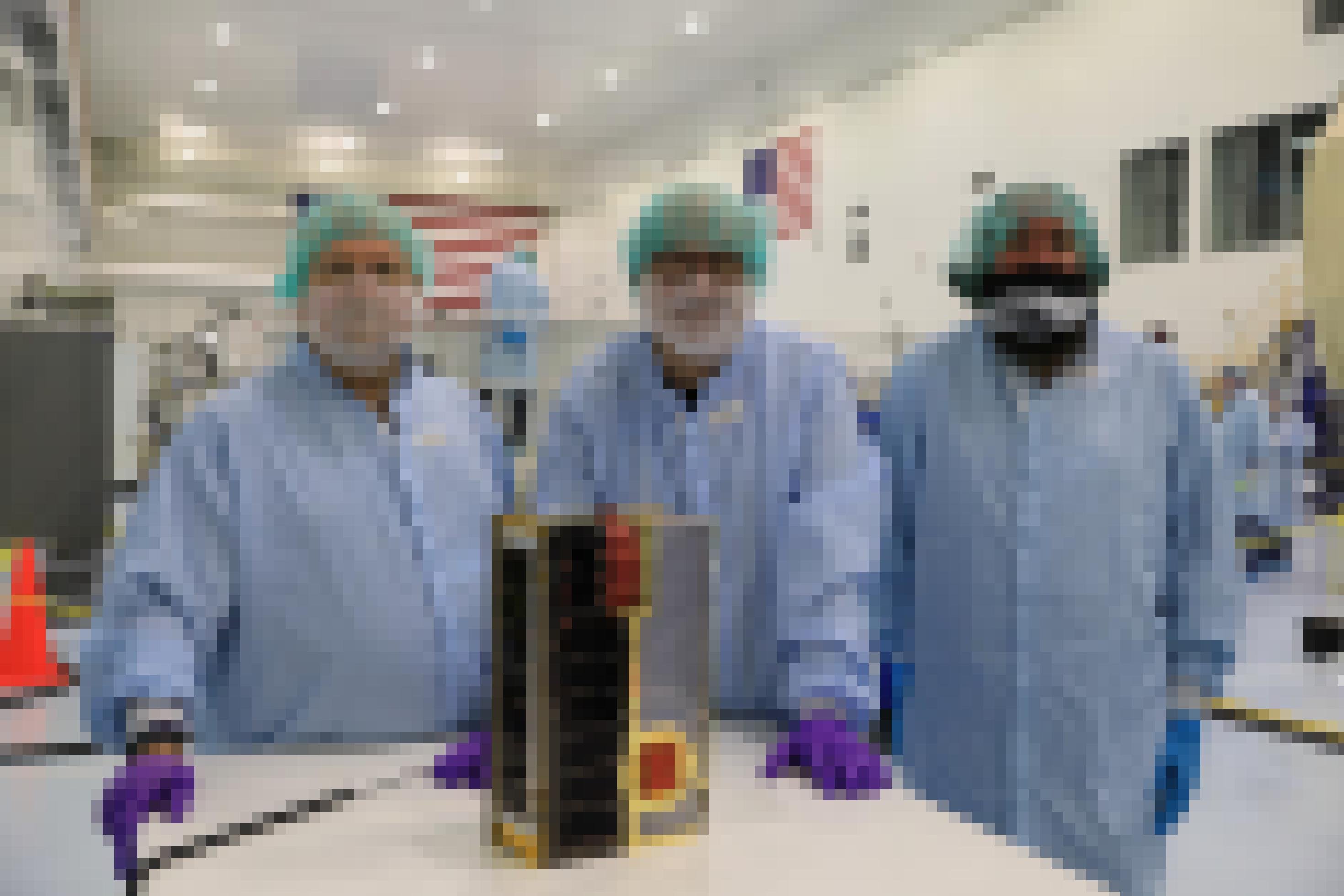 Das Team, bestehend aus drei Männern in Laborkleidung und Maske, der CuSP-Mission hinter ihrem kleinen Cubesat. Der Satellit ist ungefähr so groß wie ein Schuhkarton. Sie befinden sich in einem Labor.