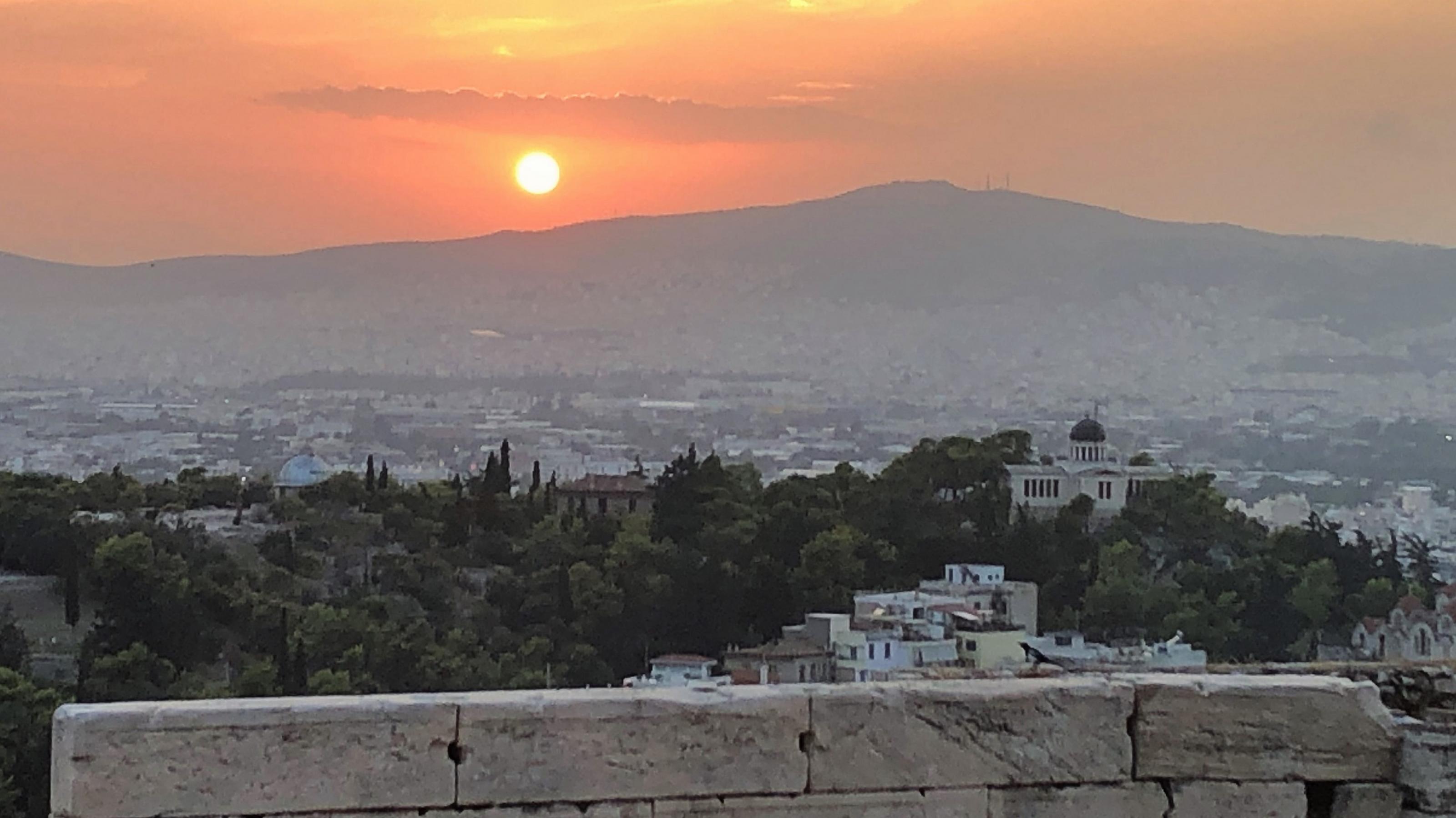 Die glühend orangene Sonne verschwindet hinter einem Hügel. Darunter das endlose Häusermeer Athens. Im Vordergrund eine Mauer der Akropolis.