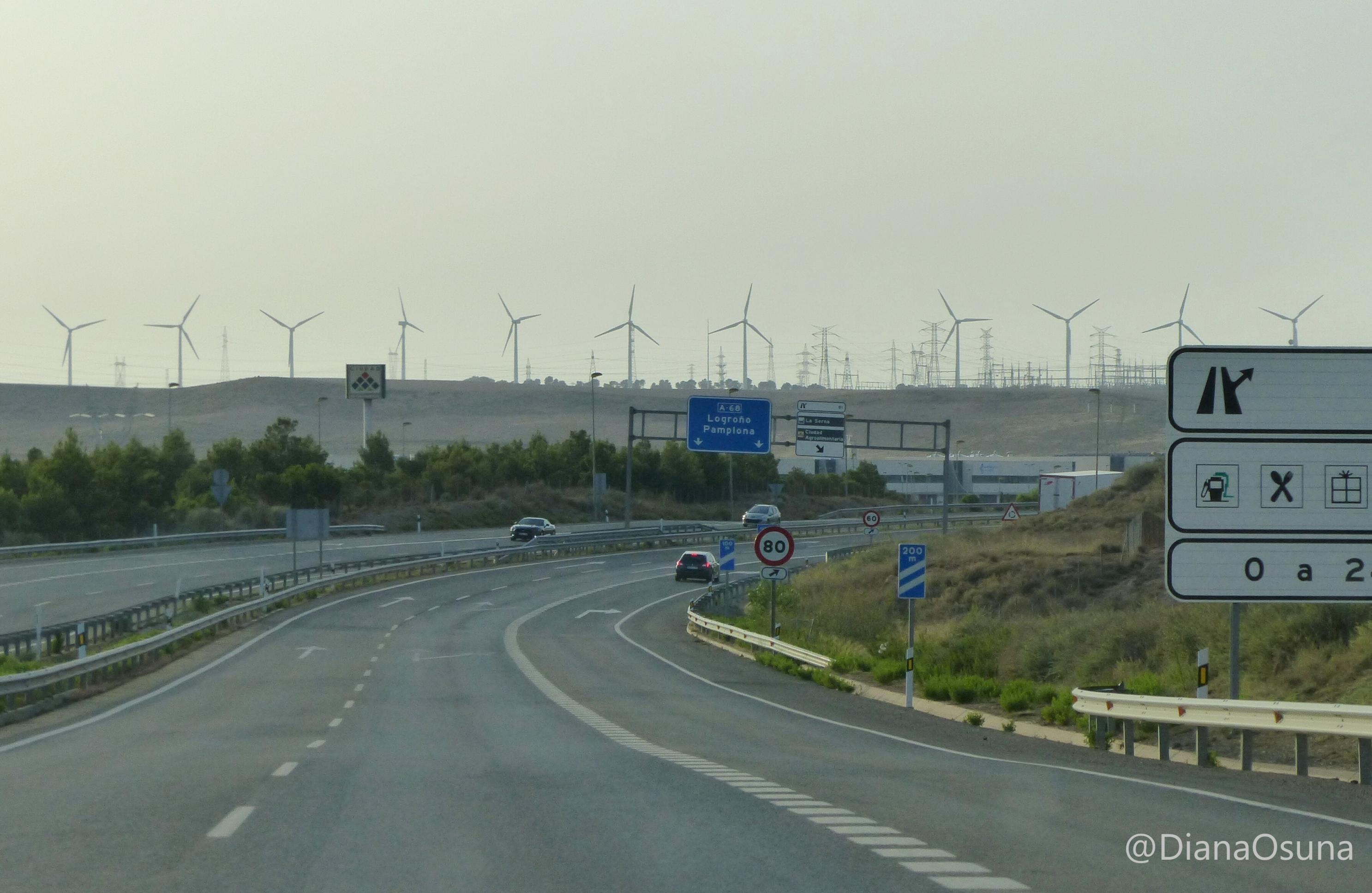Am Horizont hinter einer Autobahnausfahrt steht eine lange Reihe von Windrädern und Strommasten