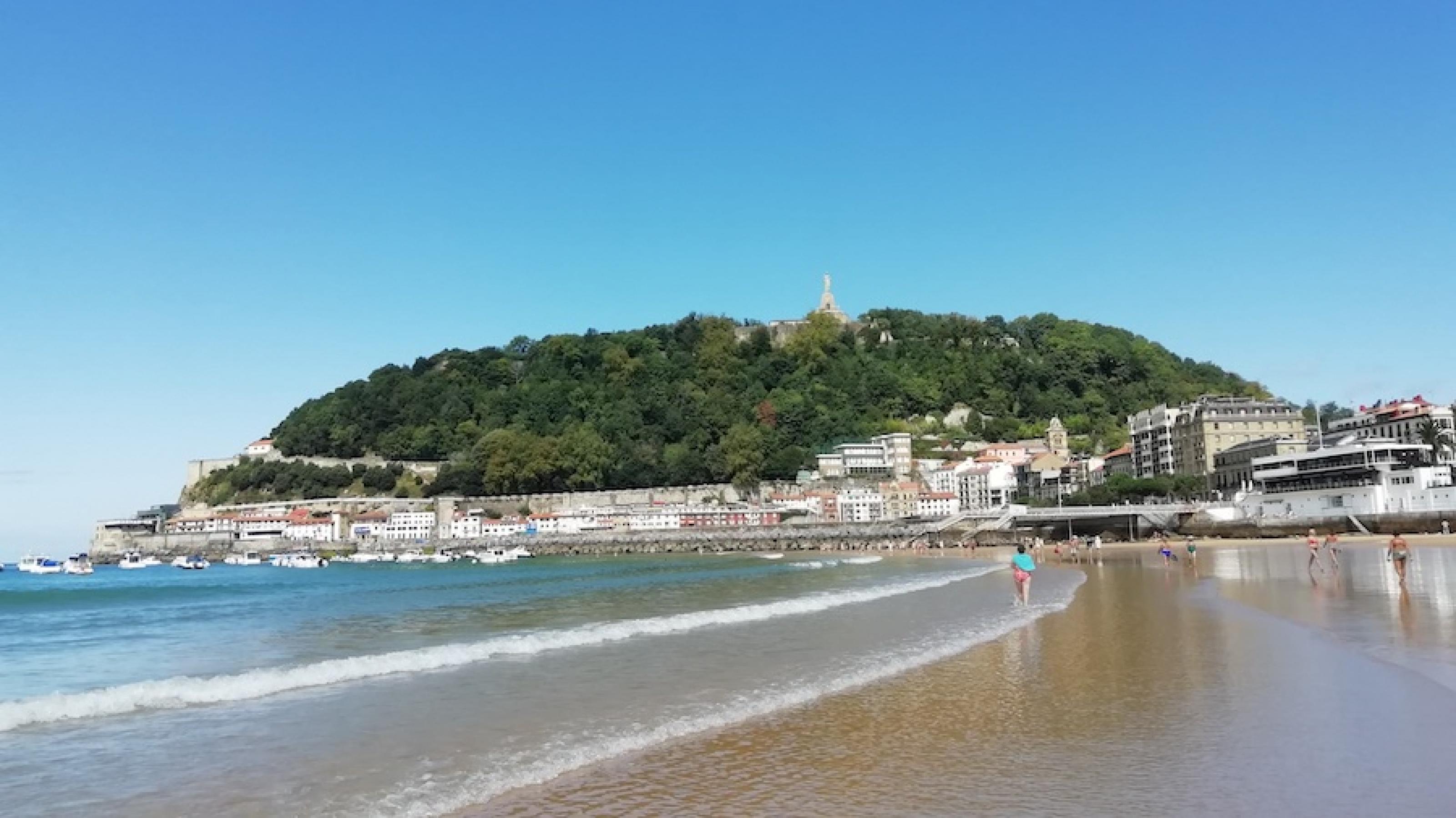 Viele Menschen gehen am Strand von San Sebastian spazieren, im Hintergrund sind große Gebäude und Boote vor einem grün bewachsenen Hügel zu sehen.