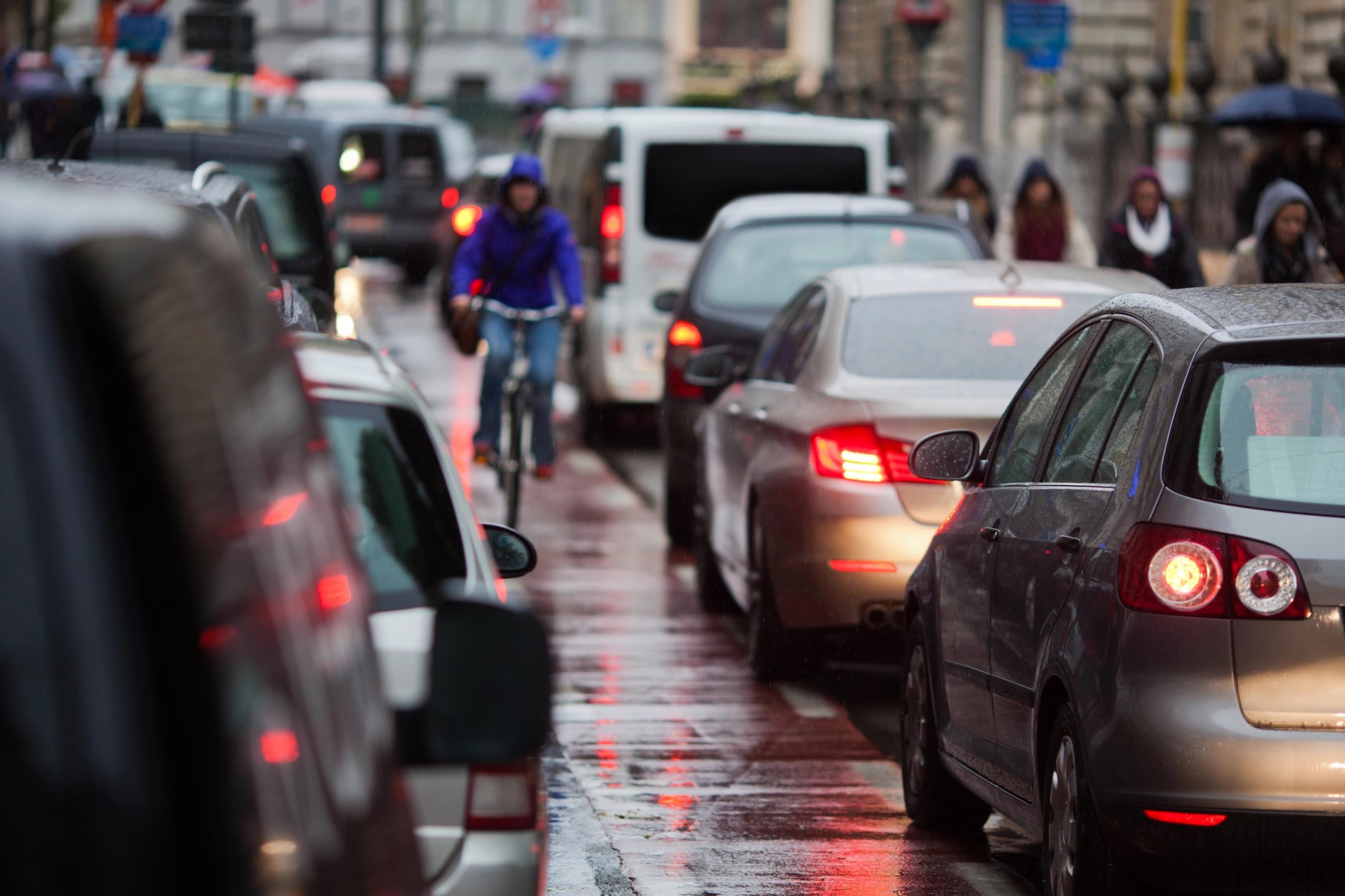 Ein Radverkehr fährt in der Mitte der Fahrbahn auf einem markierten Radweg. Rechts und links von ihm fahren Autos. Es regnet.