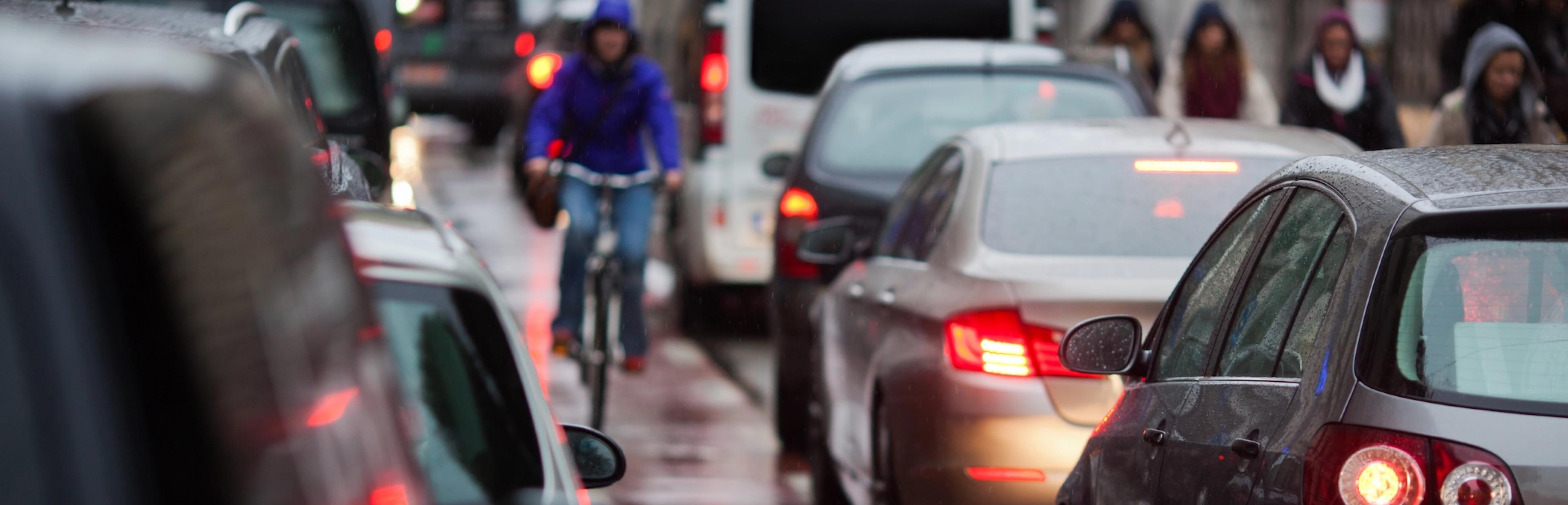 Ein Radverkehr fährt in der Mitte der Fahrbahn auf einem markierten Radweg. Rechts und links von ihm fahren Autos. Es regnet.