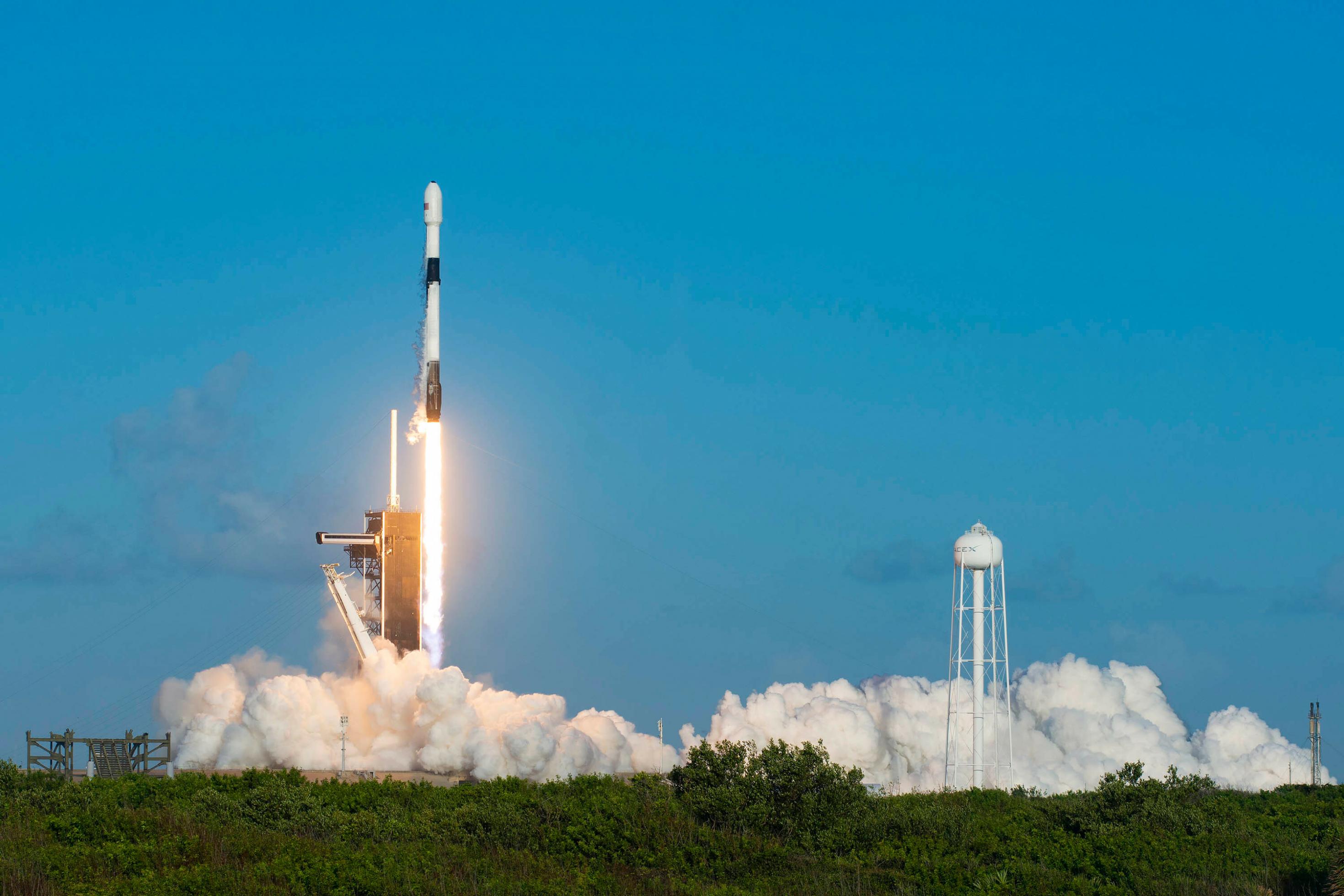 Raketenstart vor grünem Buschland, rechts davon ein Wasserturm