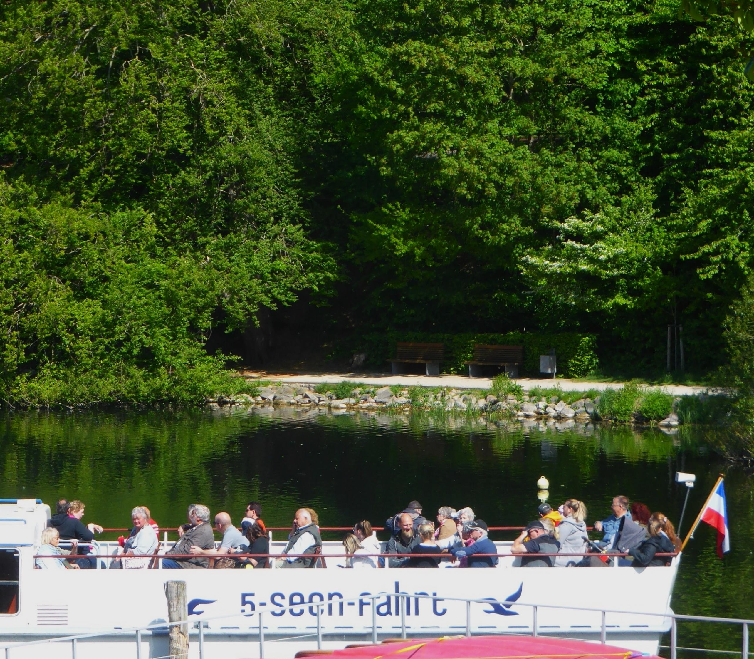 Im hinteren, offenen Teil eines weißen, mit der Aufschrift 5-Seen-Fahrt markierten Schiffs, wartet eine im Schnitt eher betagte Gästeschar sitzend auf die Abfahrt über den Dieksee, hinter dem eine sattgrüne „Wald-Wand“ aufzuragen scheint.