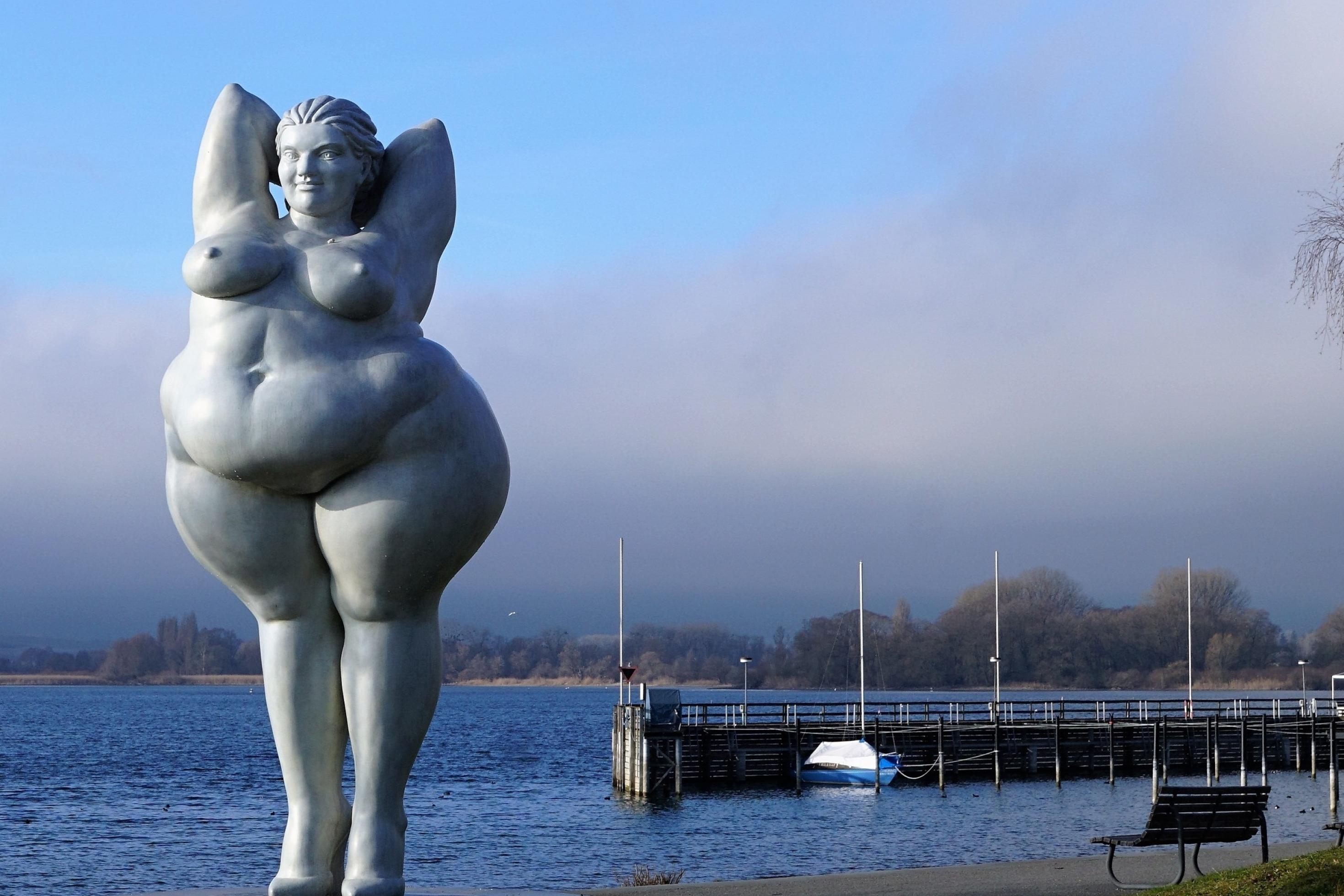 Vor dem Hintergrund des Bodensees steht auf einer Wiese die ästhetische Statue einer dicken Frau.