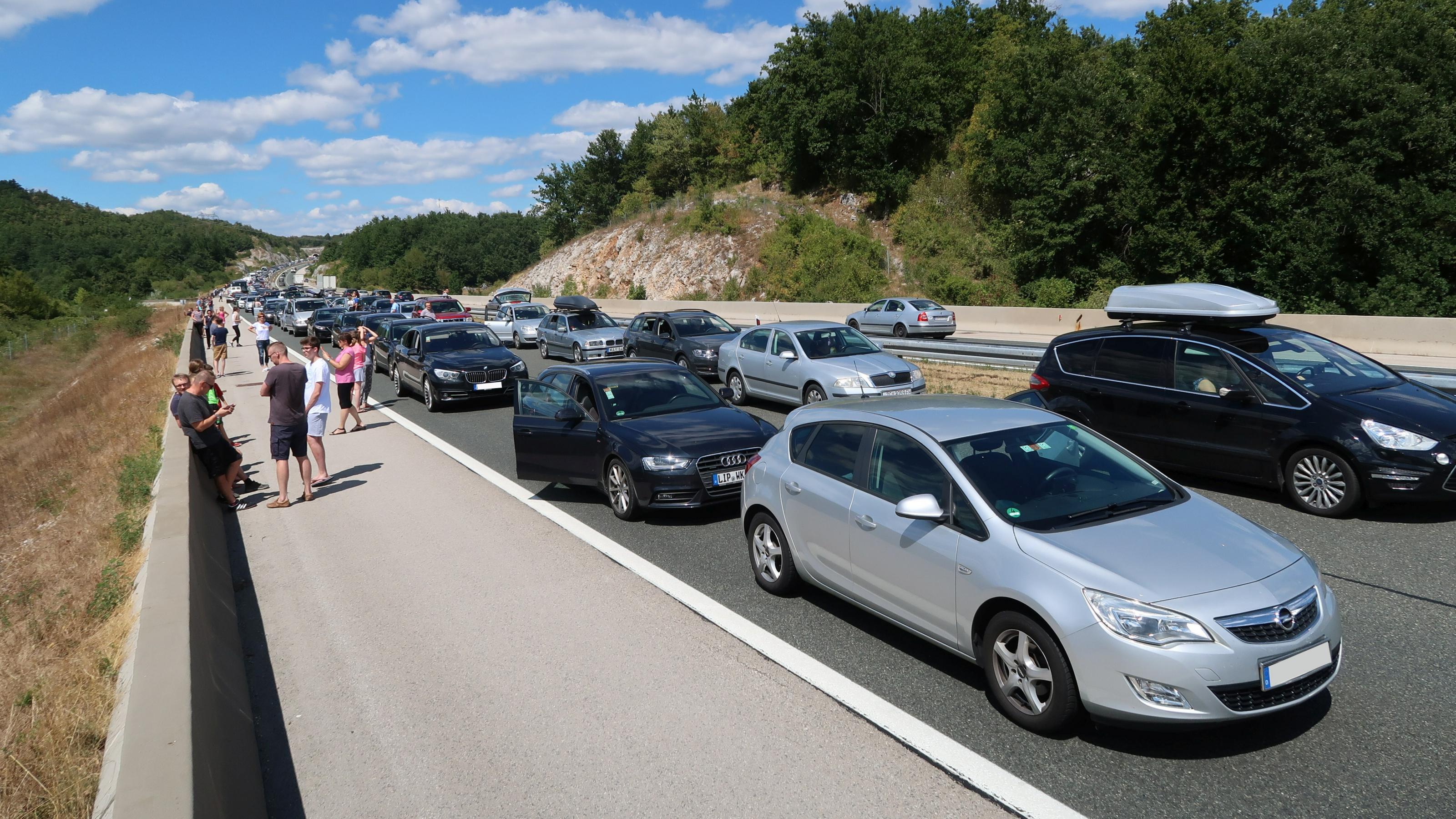 Auf einer Autobahn ist ein langer Stau zu sehen. Mehrere Personen stehen neben ihren Autos und unterhalten sich.