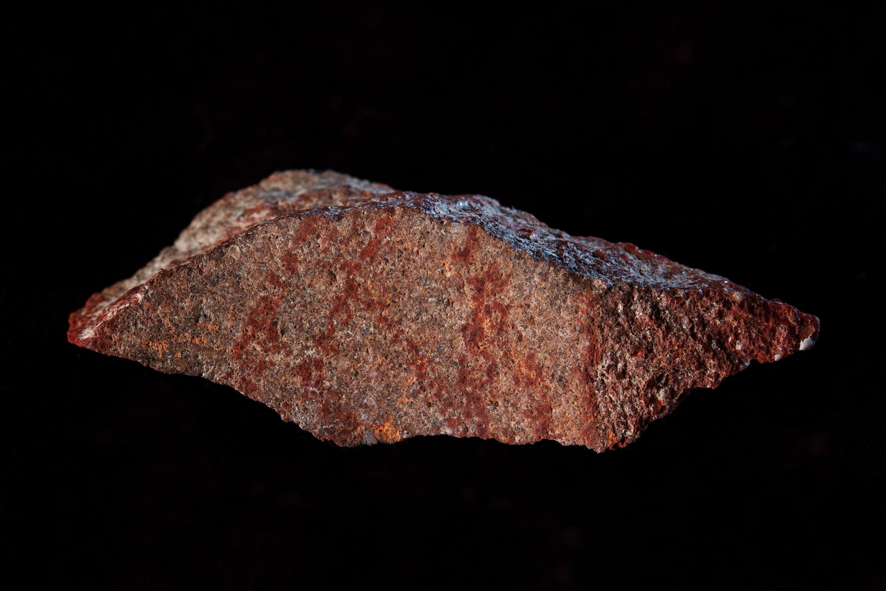 Vor schwarzem Hintergrund ist ein länglicher, rotbrauner Stein mit scharfen Kanten und einer relativ ebenen Vorderseite zu sehen. Auf ihr ist ein dunkelrotes, abstraktes Muster aus mehreren Strichen zu erkennen.