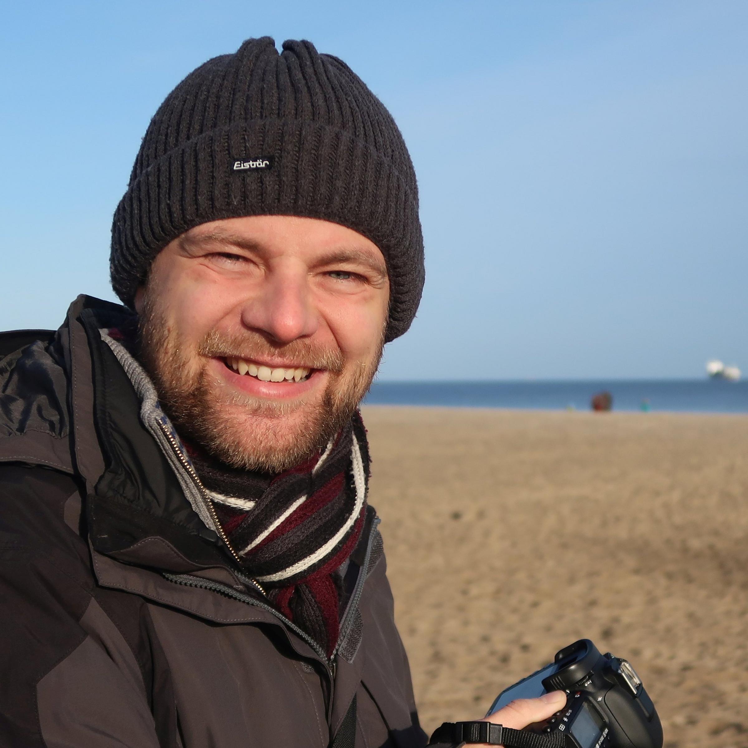 Mann mit Mütze und Winterjacke steht am Strand und hält Kamera in der Hand.