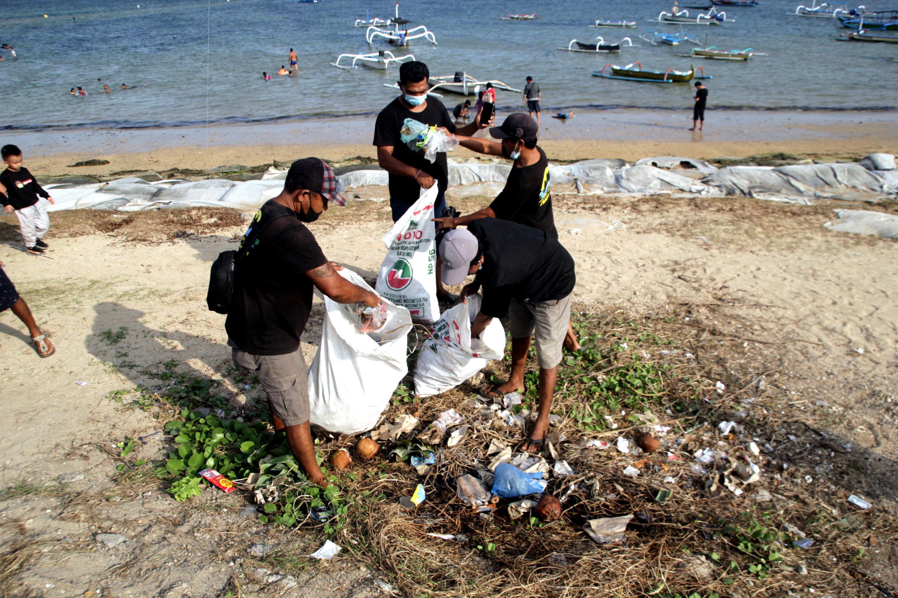 Drei Personen stehen vor dem Meer an einem Strand mit großen weißen Plastiktüten und füllen sie mit Müll, der auf dem Boden liegt.