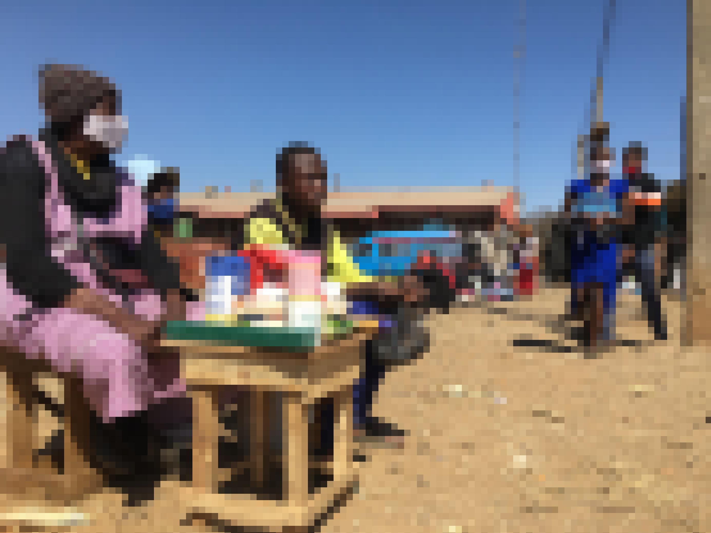 Das Bild zeigt die Straßenhänderlin Vangayi Maisiri, die hinter ihrem kleinen Stand sitzt, auf dem die Waren aufgestellt sind. Im Hintergrund ein paar Passanten.