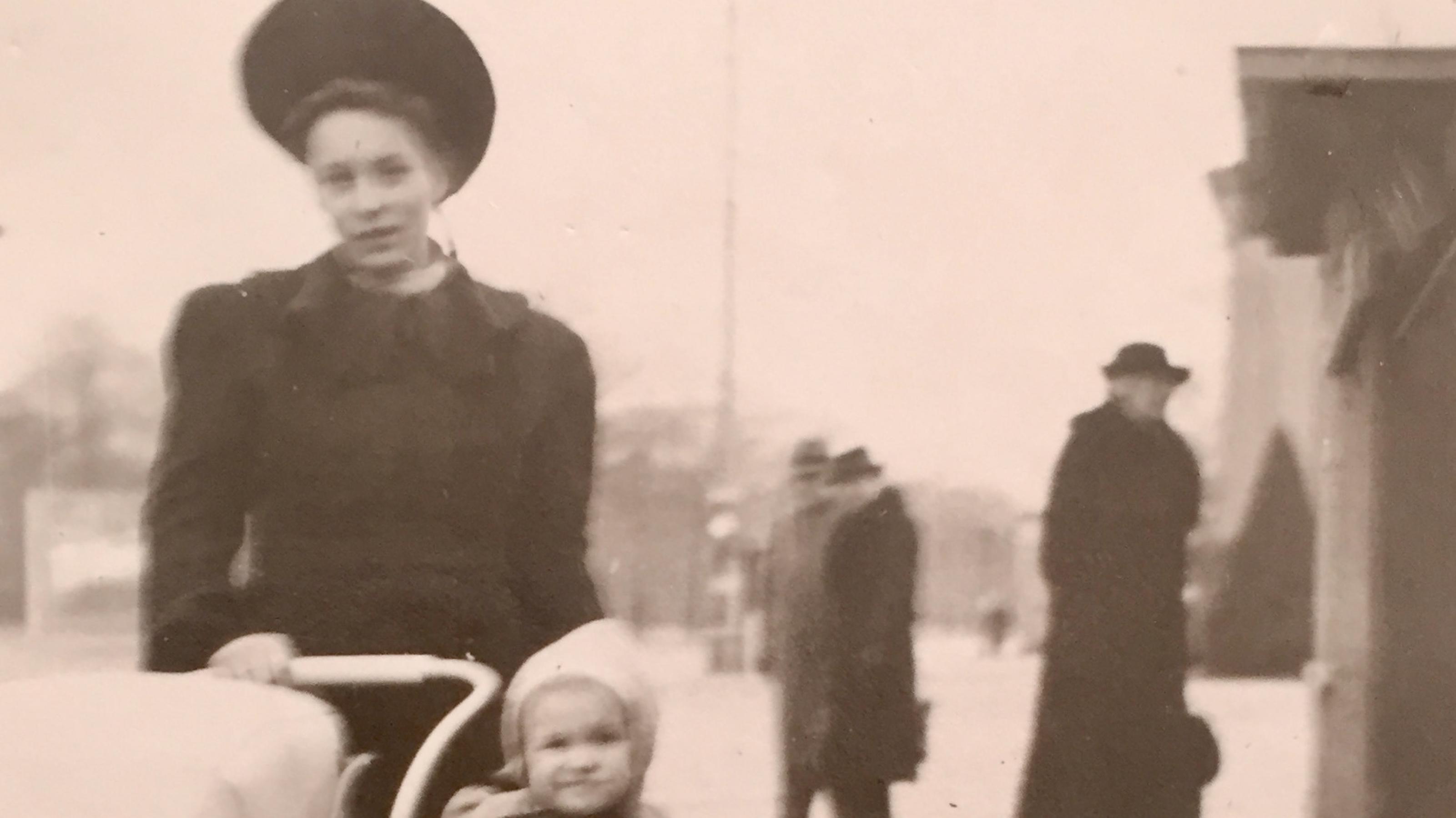 ein schwarz-weiss Foto von Angela, ihrer Mutter und kleinen Schwester 1945, Strassenszene in Dresden vor der Bombardierung, eine Frau mit Hut schiebt einen Kinderwagen, neben ihr ein kleines Kind mit weißer Wollmütze. Frau und Kind lächeln in die Kamera.