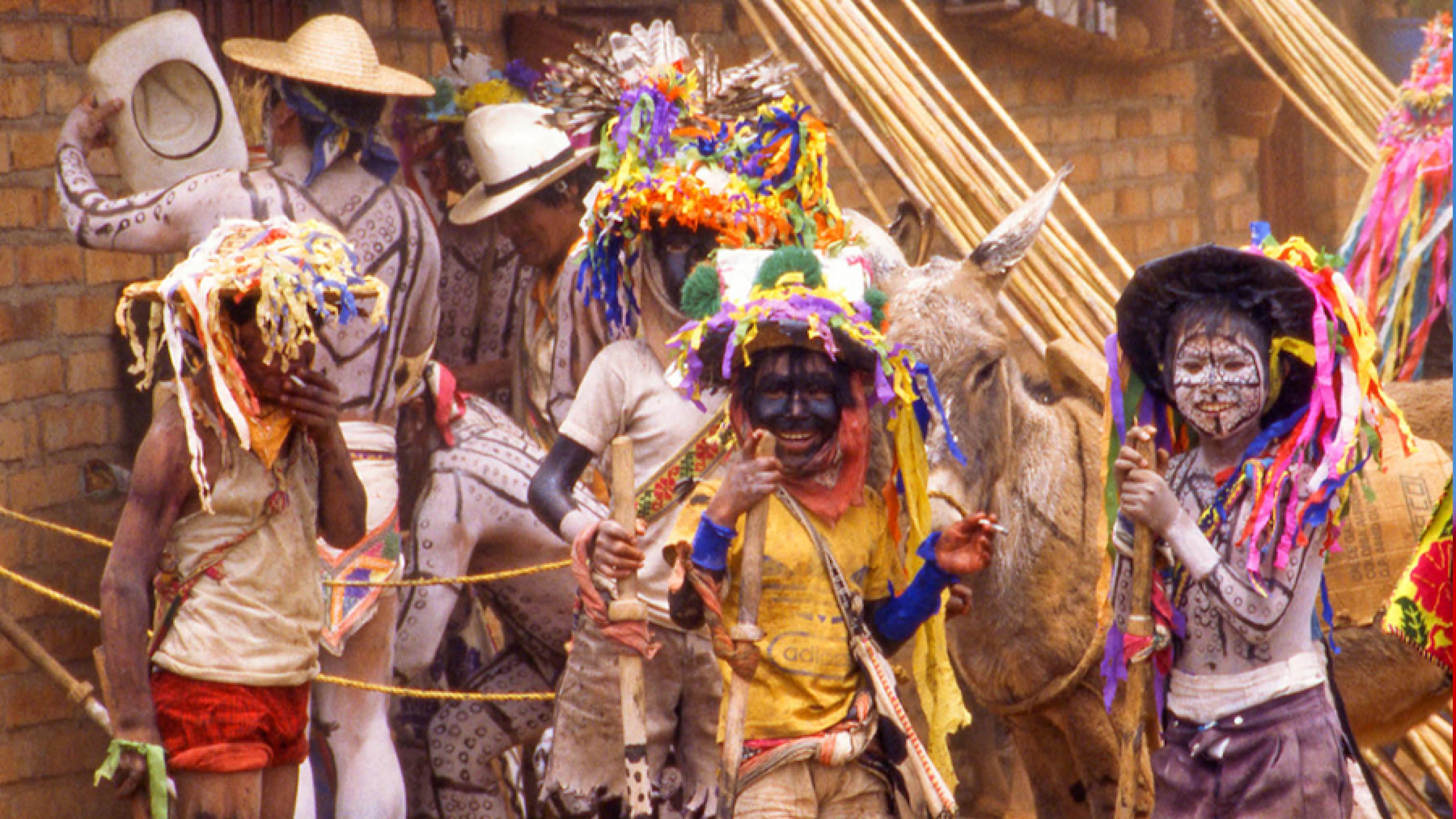 Straßenszene aus Mexiko City mit bunt gekleideten Feiernden. Dazwischen Esel und Sombrero-Verkäufer