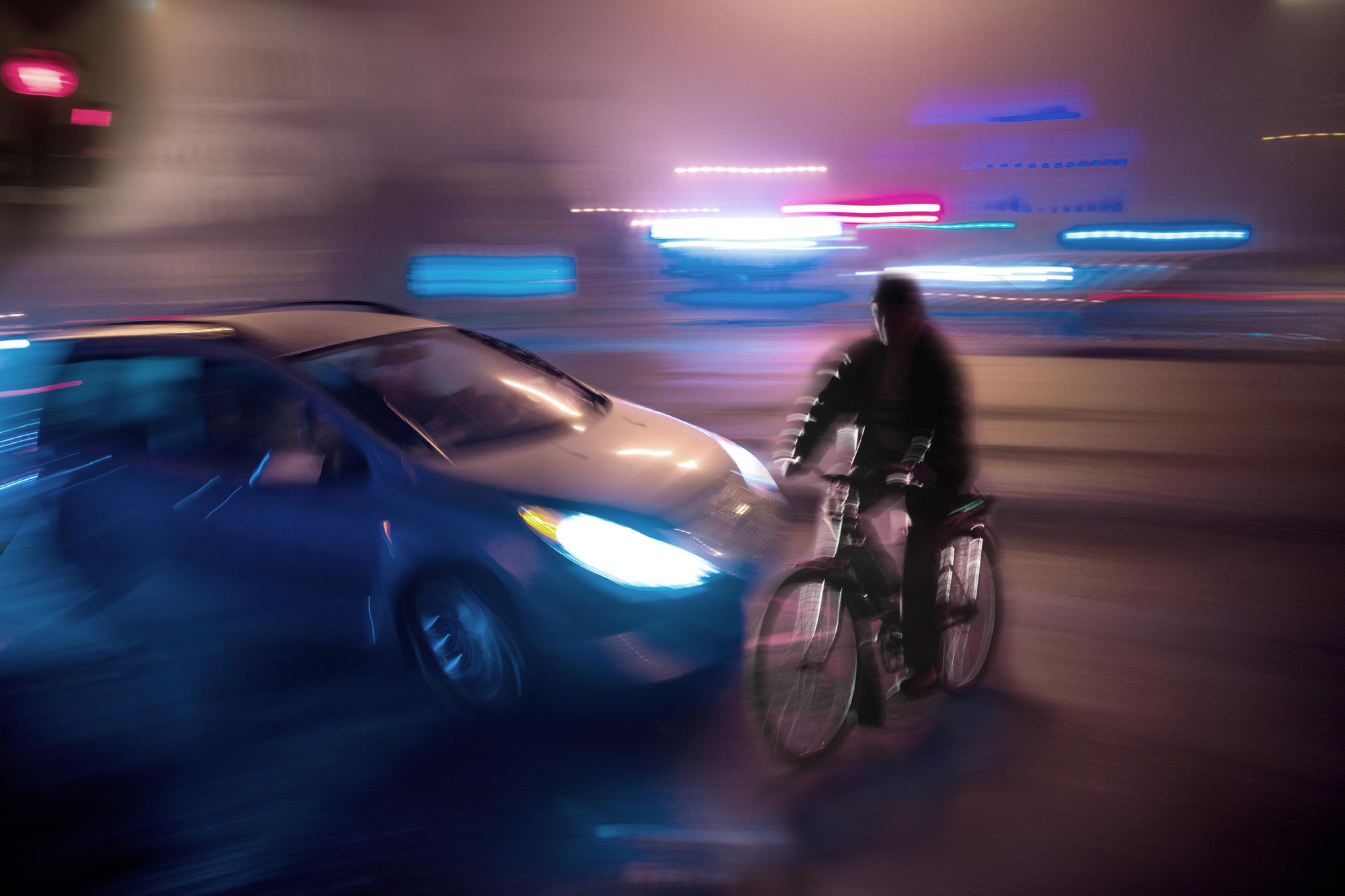 Bei Nacht: Ein Auto schneidet knapp einen Fahrradfahrer, der die Straße überquert. Die Straße ist im Vordergrund dunkel, im Hintergrund leuchten hellblaue und rote Lichter im Strobo-Effekt.