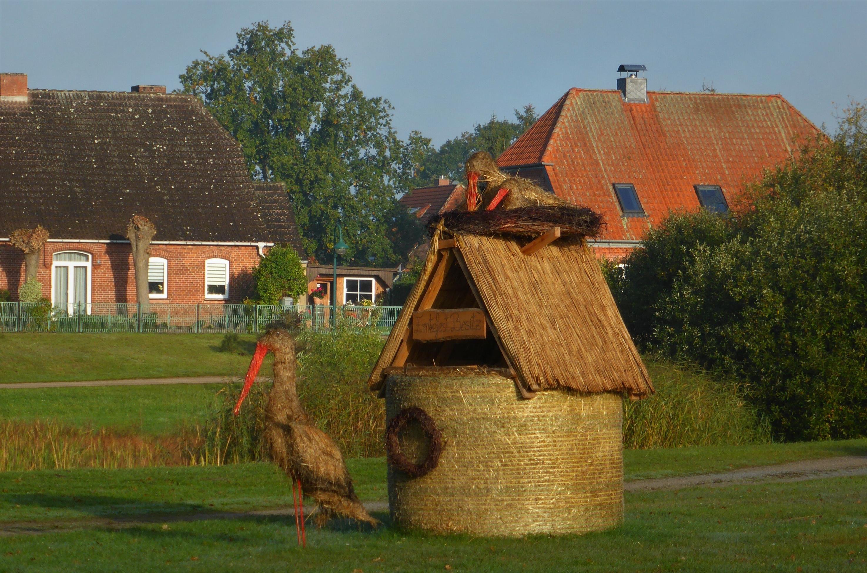 Vor zwei Rotklinker-Dorfhäusern steht ein aus Stroh und Holz gebaute Mini-Kate, Aufschrift „Erntefest Besitz“. Davor steht ein „Stroh-Storch“ und ein ebensolcher sitzt in einem Strohnest, das auf der Kate montiert ist.