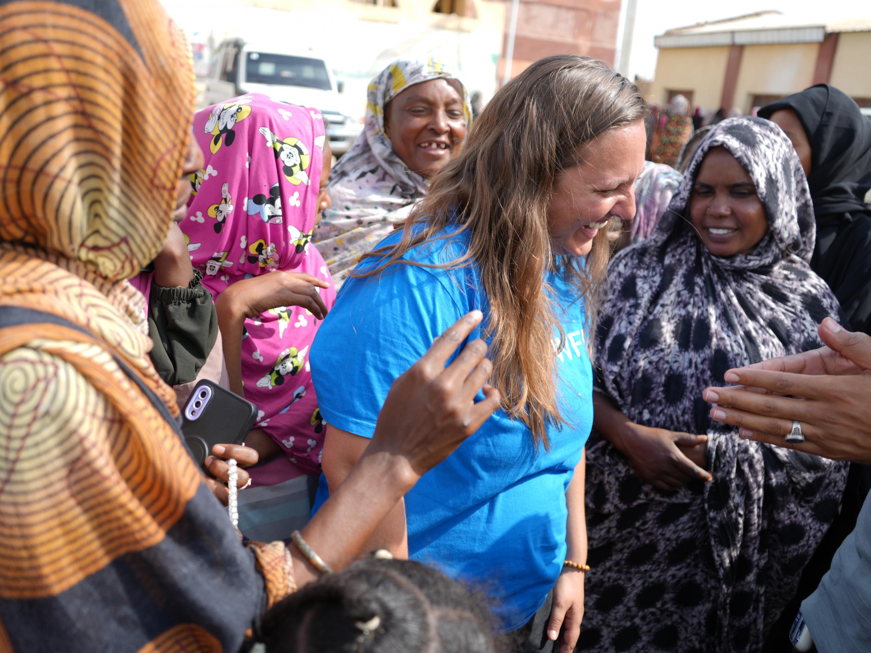 Eine junge Frau steht inmitten einer Menschenmenge. Die Gesichter sind freundlich. Die Frau hat lange blonde Haare und trägt ein leuchtend blaues T-Shirt mit dem Aufdruck des WFP, die Frauen um sie herum tragen bunte lange Gewänder und haben ihre Haare bedeckt.