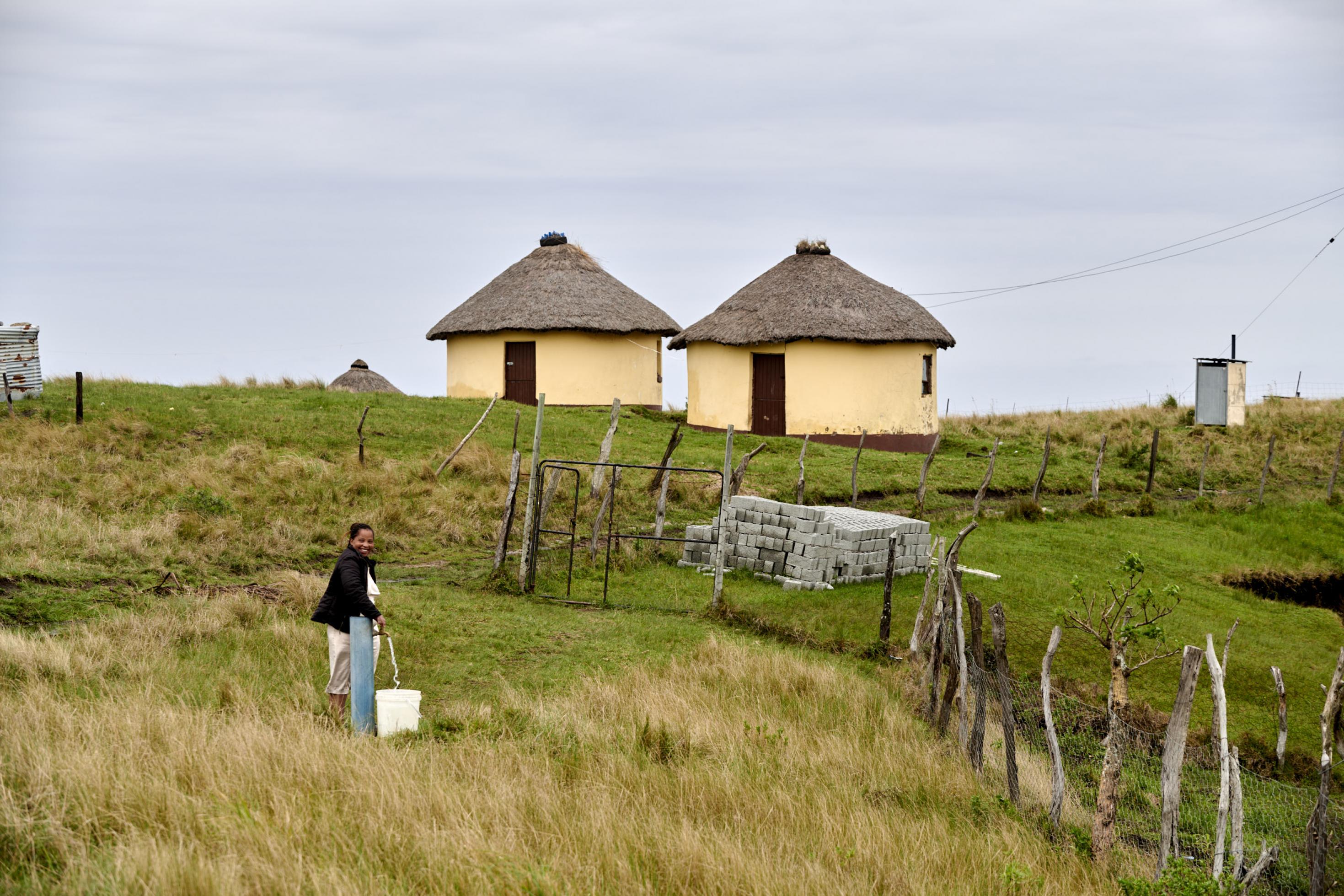 Vor zwei Rundhäusern im Dorf füllt eine Frau Wasser aus dem kommunalen Hahn in einen Eimer