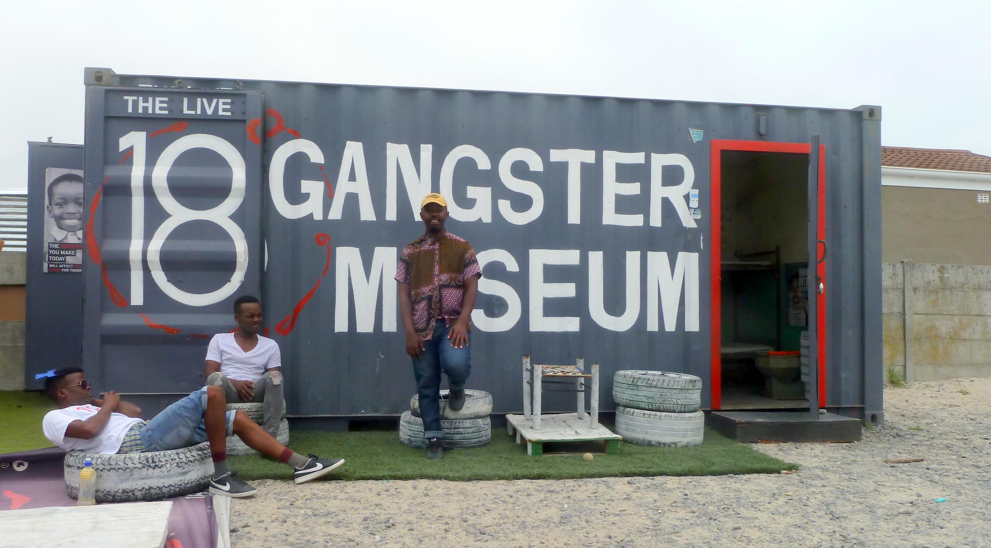 Das Foto zeigt den Museumsgründer, der an dem Schiffscontainer lehnt, das als Museum dient, daneben sitzen seine Mitarbeiter, zwei Ex-Gangster.