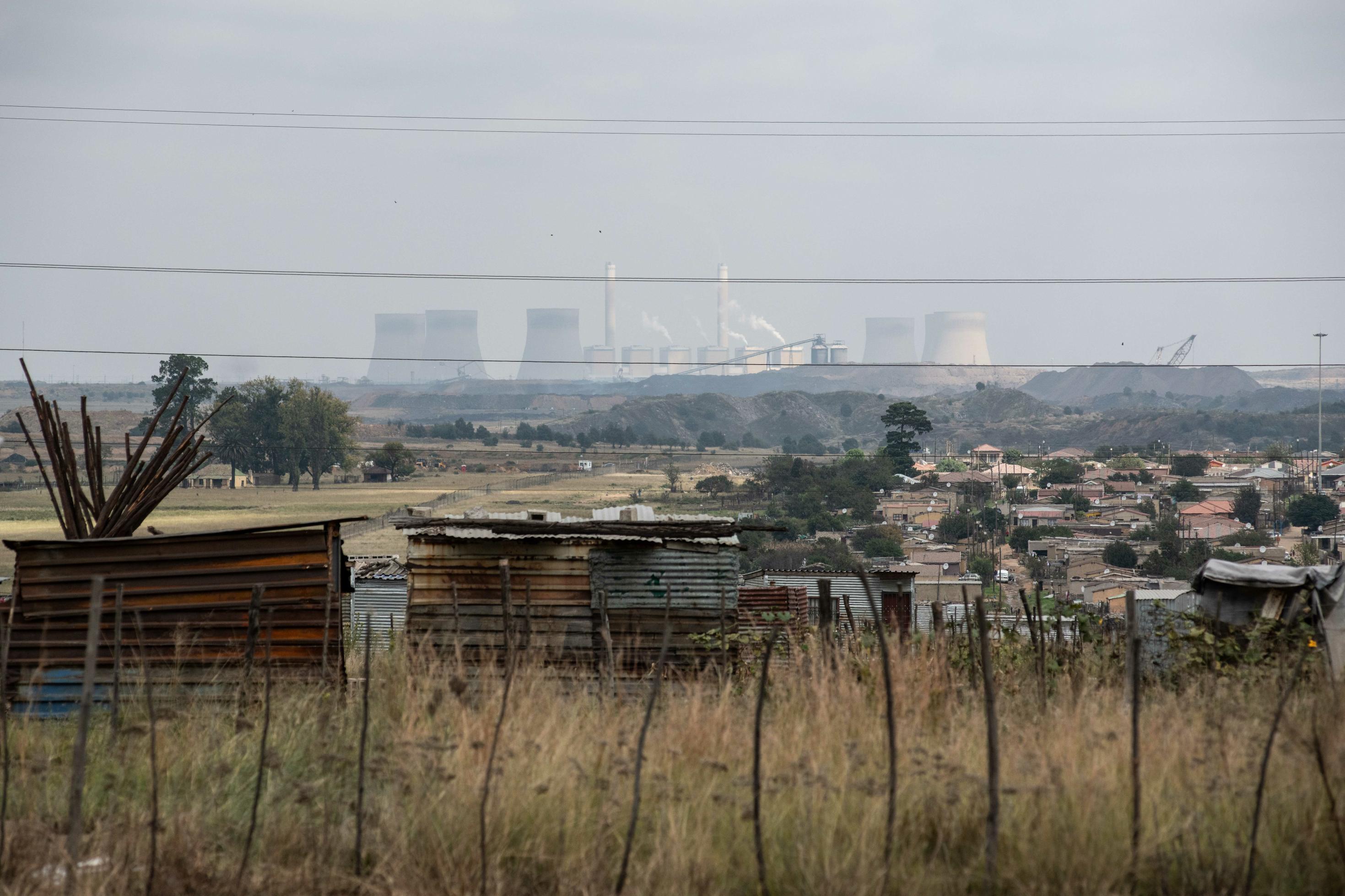 Hinter einer Siedlung kleiner Häuser sieht man ein Kohlekraftwerk am Horizont, das toxische Abgase in die Luft bläst.