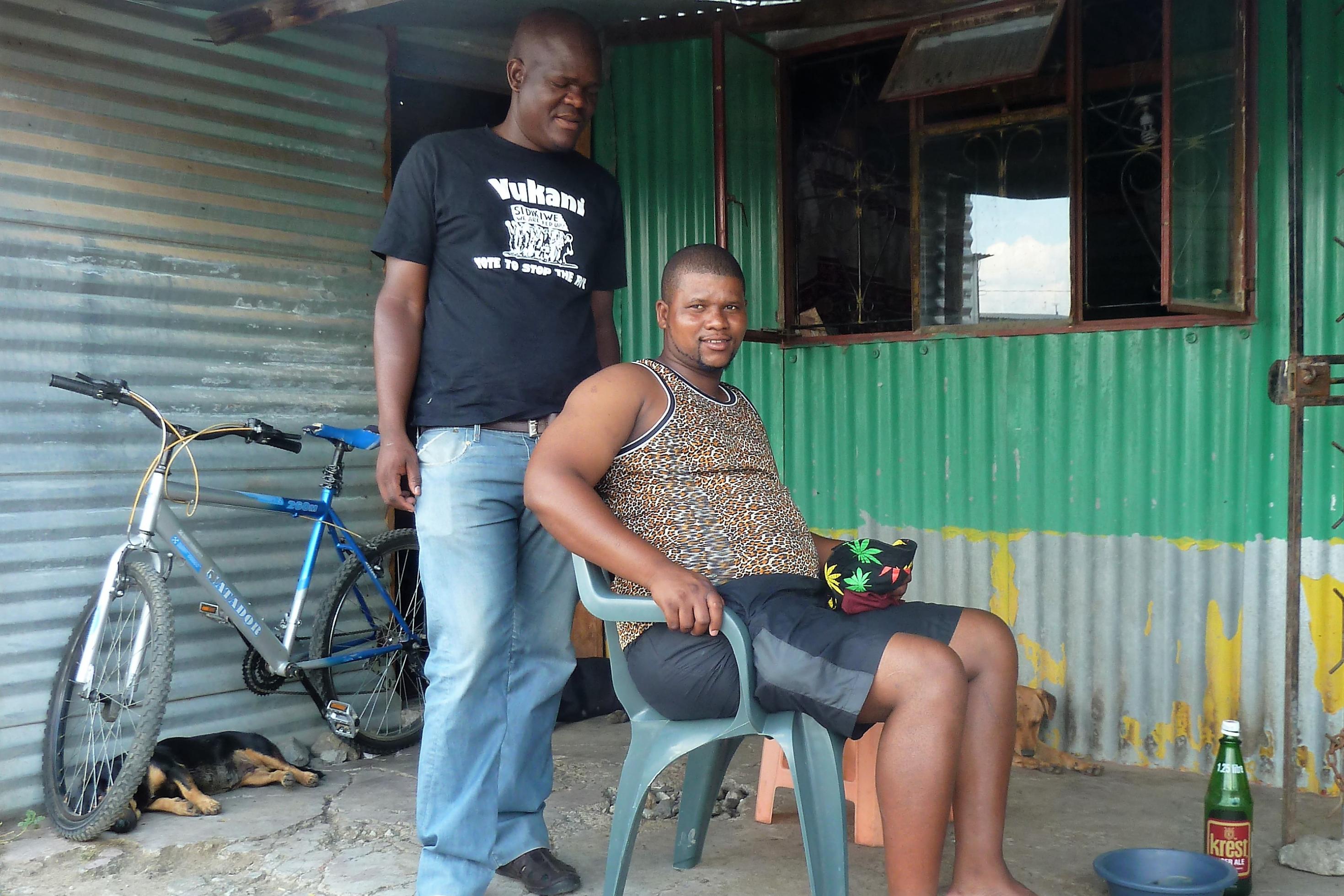 Magidiwana sitzt auf einem Plastikstuhl vor einem Wellblechhaus, an dem ein Fahrrad lehnt, hinter ihm steht ein Freund von ihm.