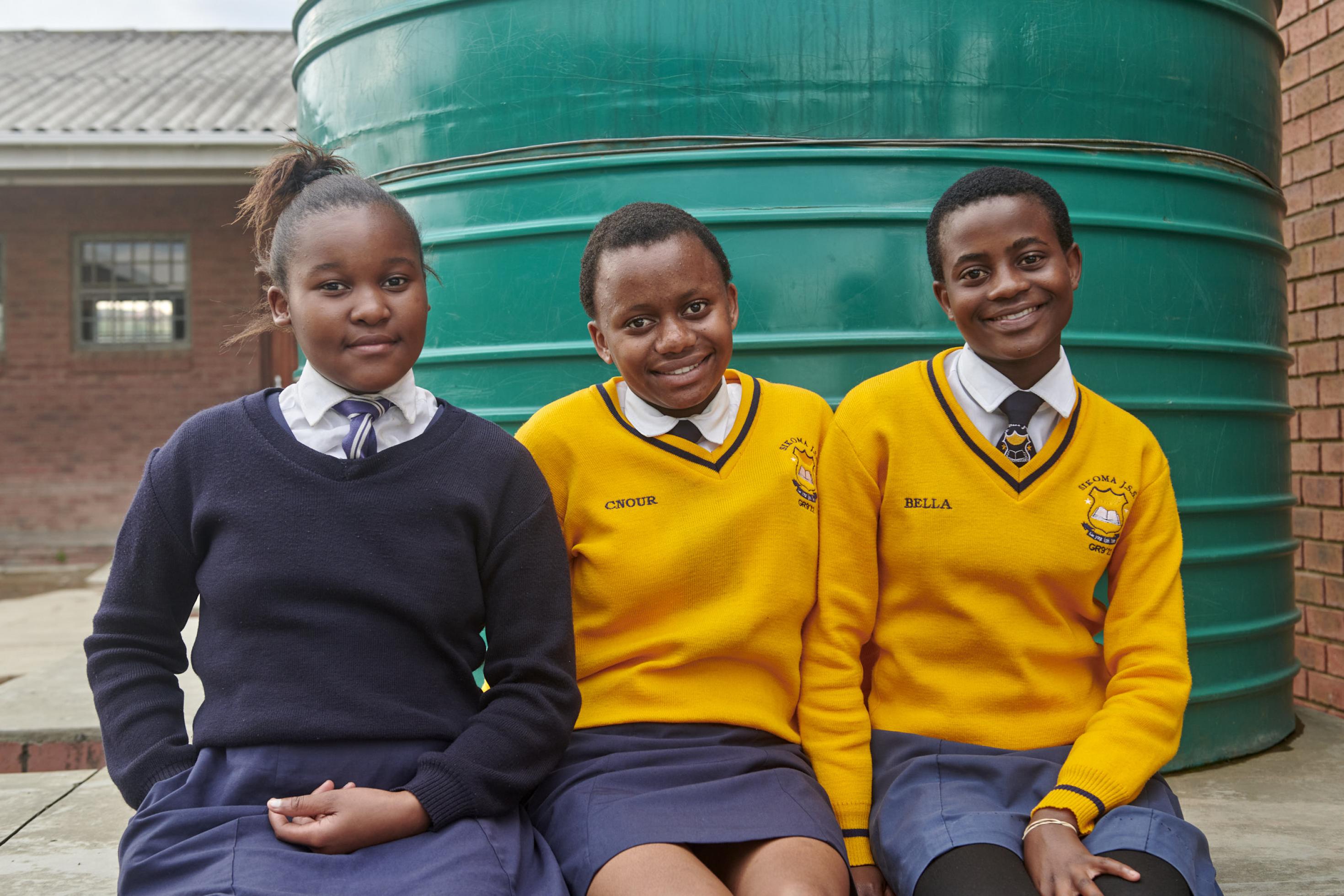 Die drei Schülerinnen sitzen vor einem Wassertank ihrer Schule, sie tragen Schuluniformen