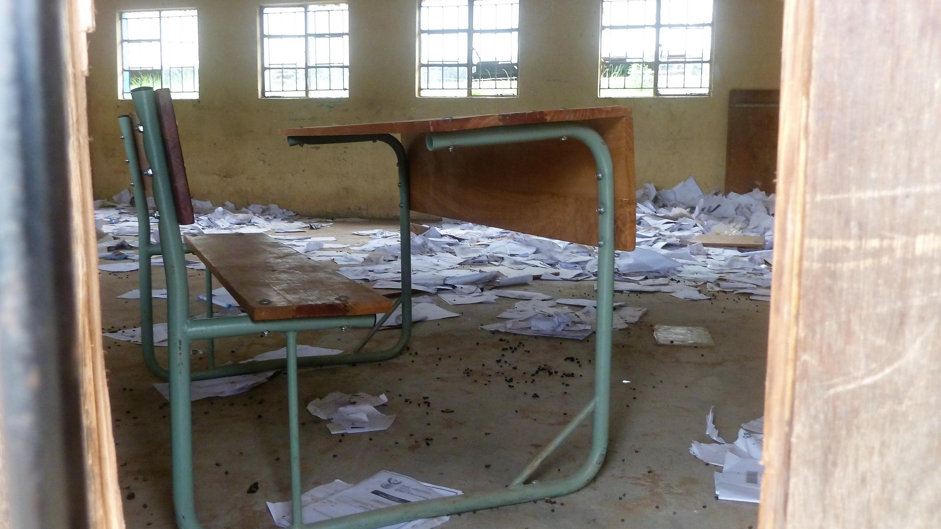 Blick durch den Türrahmen, ein Pult steht in einem leeren Klassenzimmer, auf dem dreckigen Boden liegen überall Papiere herum