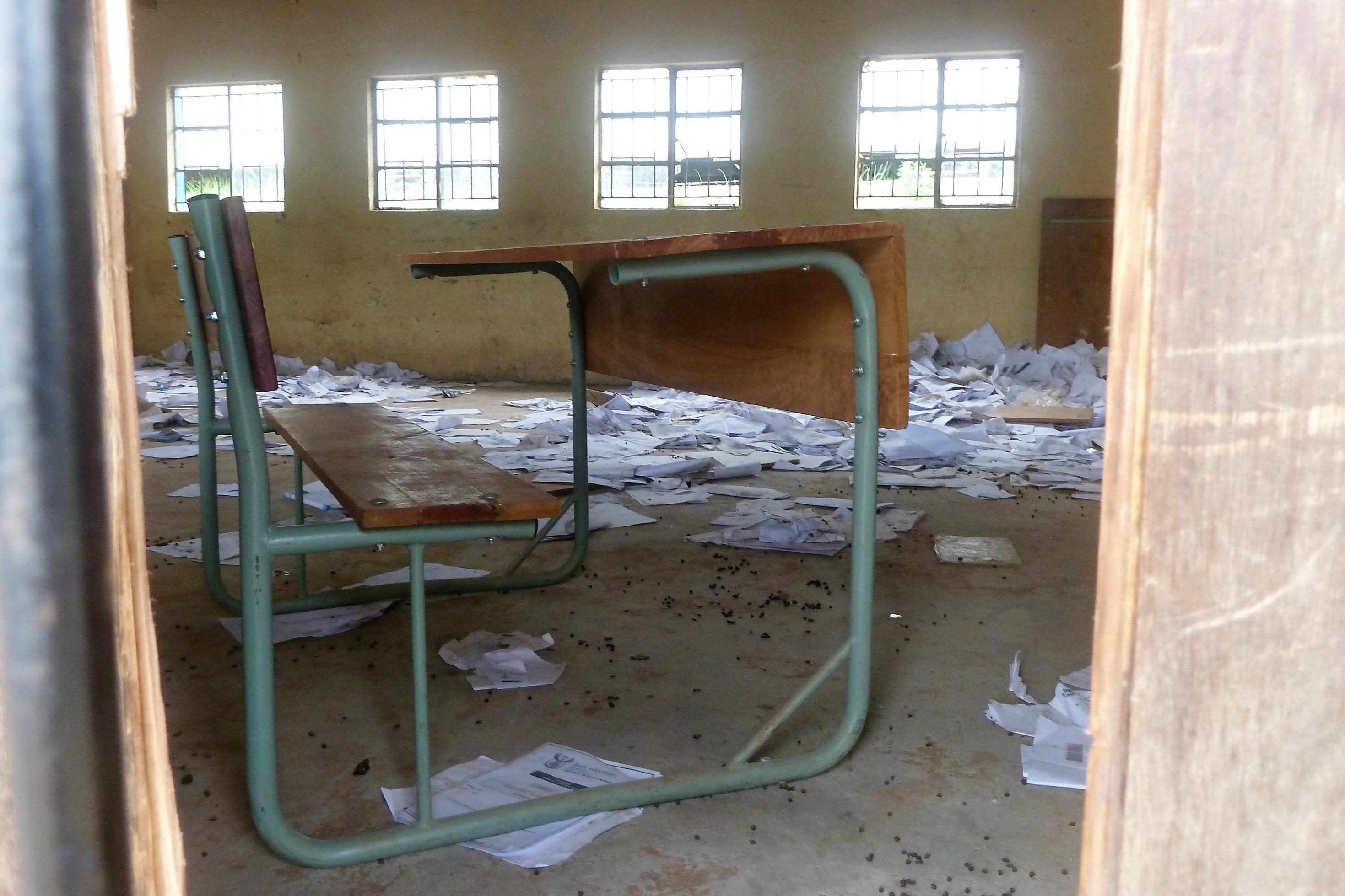 Blick durch den Türrahmen, ein Pult steht in einem leeren Klassenzimmer, auf dem dreckigen Boden liegen überall Papiere herum