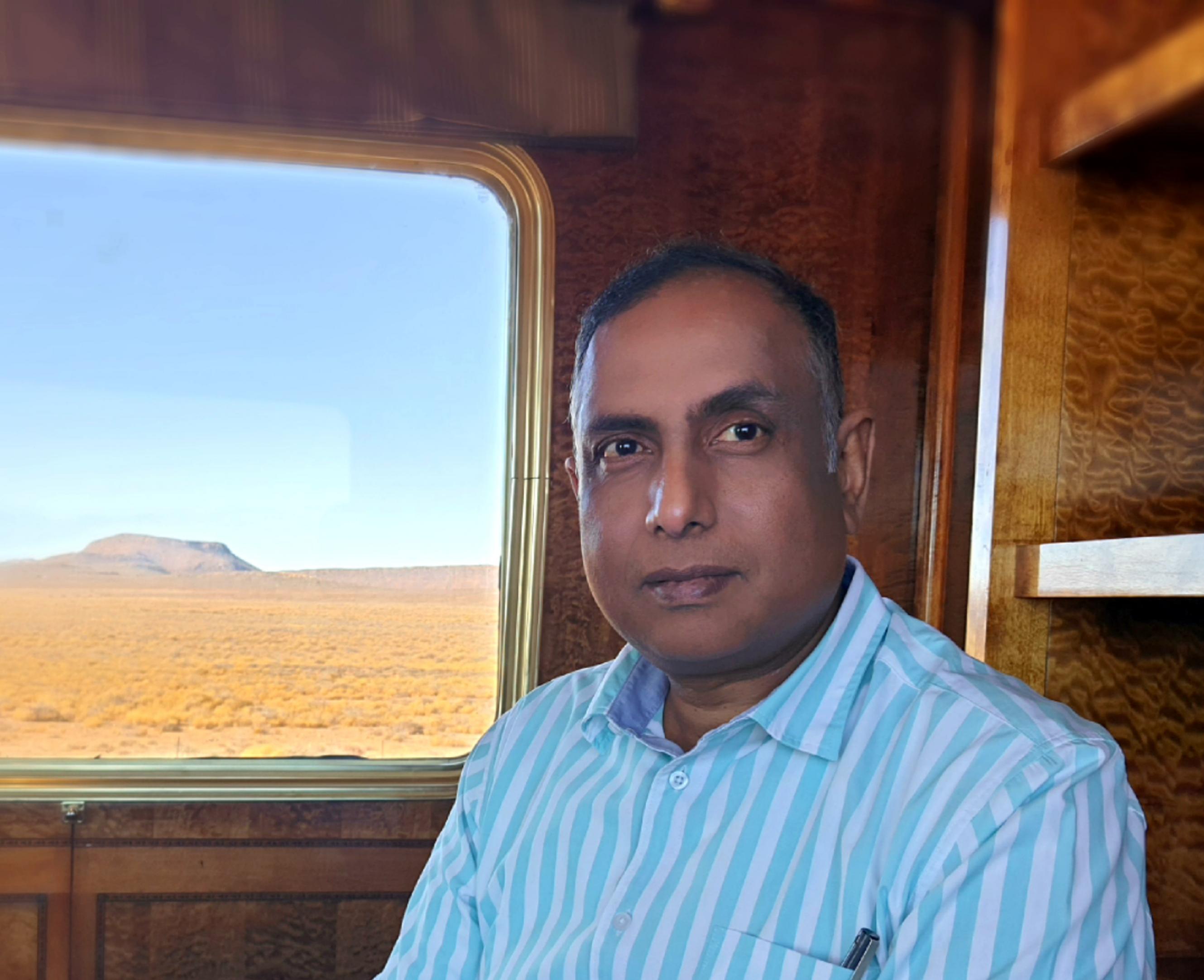 Ein Mann schräg von der Seite im Halbportrait, offenbar im Zug. In der linken Bildhälfte ist ein Fenster, der Blick geht auf eine sehr karge Landschaft, im Hintergrund ein Tafelberg.