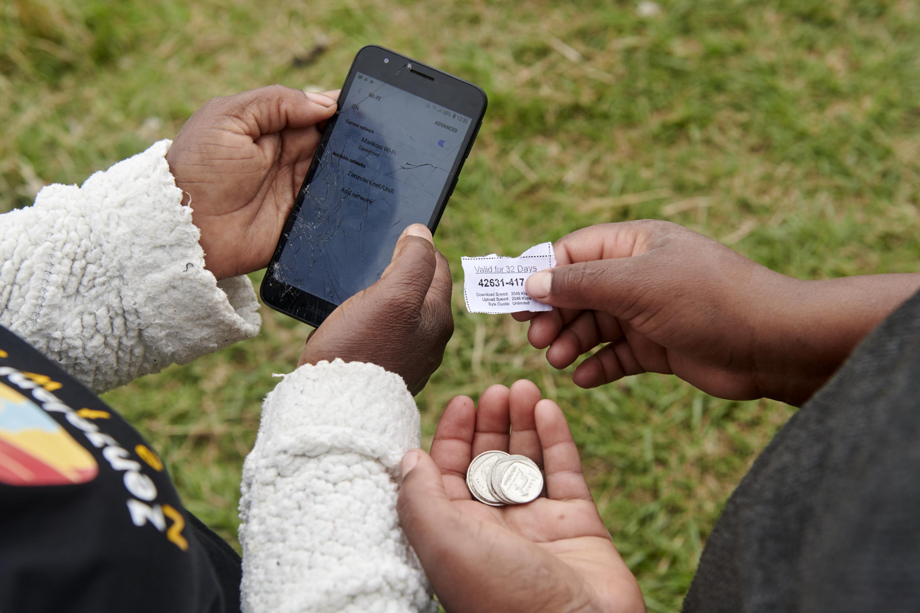 Eine Hand hält ein Smartphone, eine andere den Voucher und ein paar Münzen.