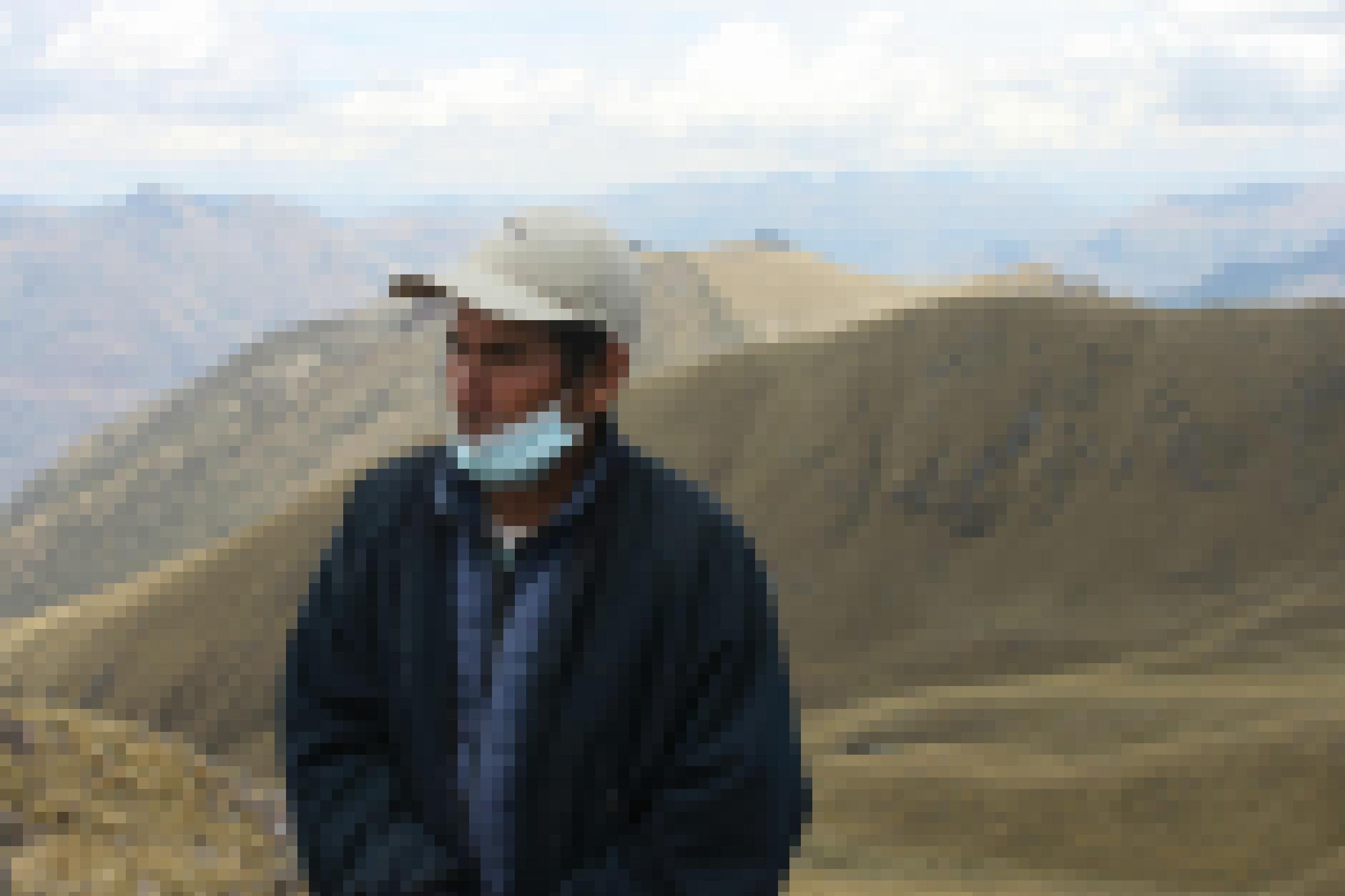 Berglandschaft, im Vordergrund ein 60-jähriger Mann mit weisser Baseballmütze und blauer Jacke, wettergegerbtem Gesicht, Mundschutz.