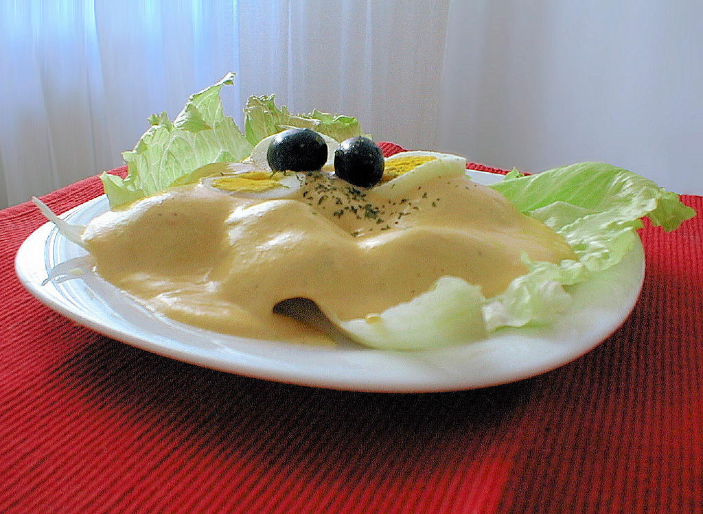 Weißer Teller, darauf Salatblätter, unter einer dicken gelben Soße sieht man die Erhebungen der darunterliegenden Kartoffelscheiben. Obenauf zwei schwarze Oliven und ein Viertel gekochtes Ei.