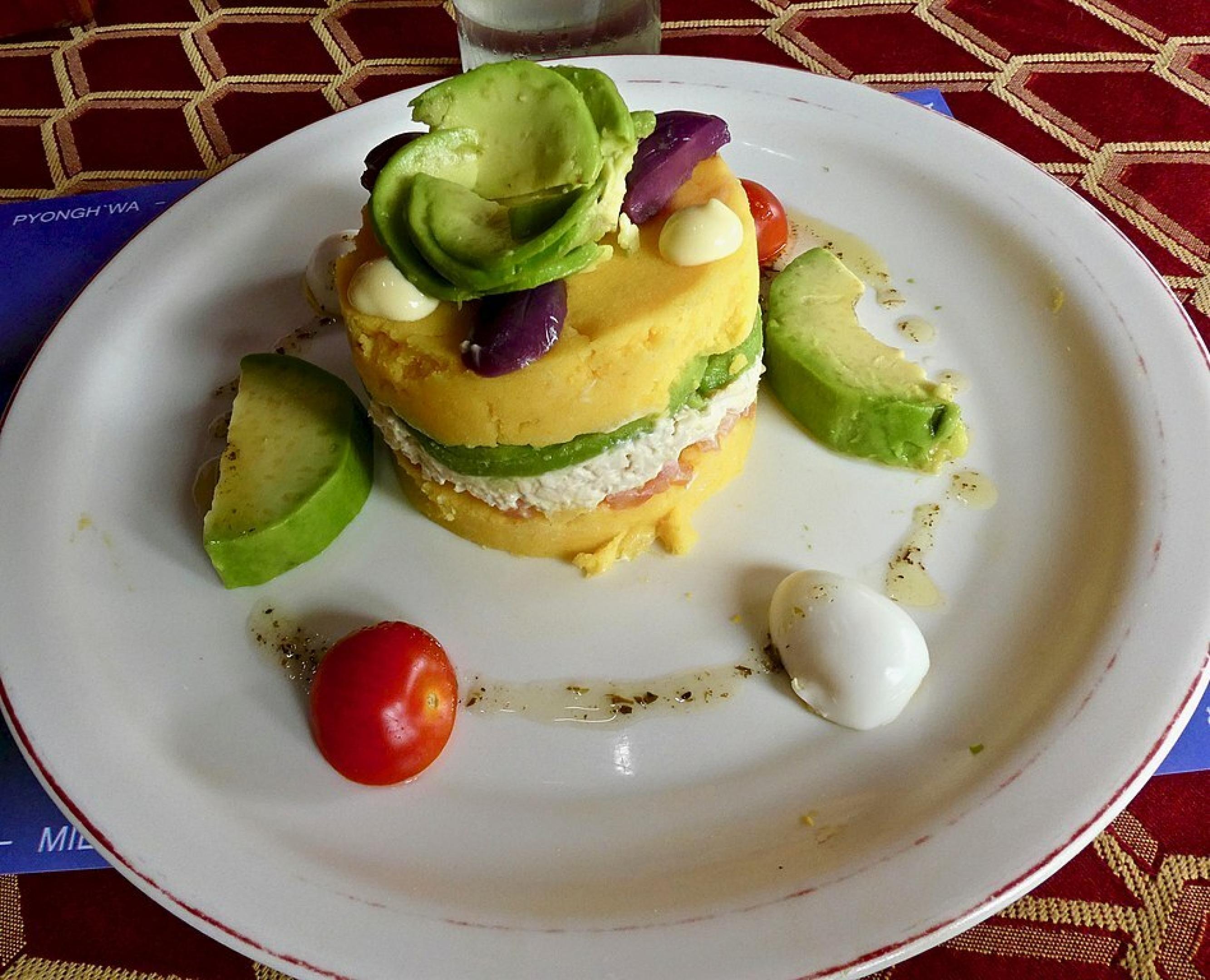 Weißer runder Teller auf einem Tisch. Darauf ein rundes Törtchen, unten gelb, dazwischen eine Schicht mit Mayonnaise und Avocado, darüber wieder gelbe Paste. Dekoriert mit einem Salatblatt, einer Olive und kleinen Stückchen gekochten Eis.